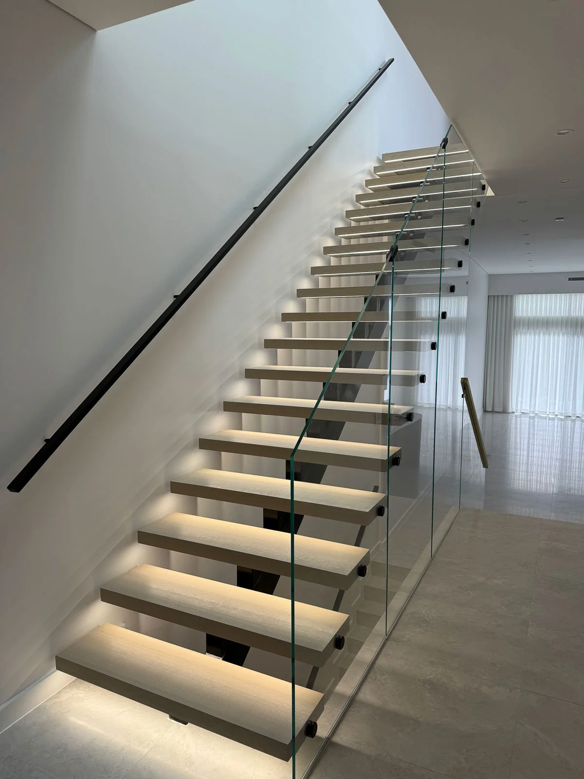 Modern interior staircase with wooden steps, glass railing, and minimal lighting in a bright, spacious room. Staircase Polishing Sydney
