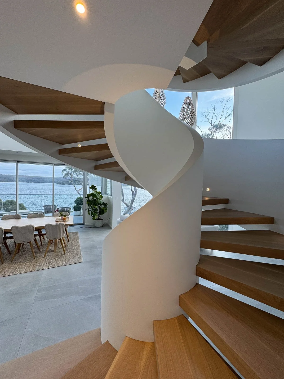 Interior view of a modern house featuring a spiral staircase with wooden steps and white railing, large windows overlooking a body of water, dining area with a long table and chairs, and decorative hanging lamps. Staircase Polishing Sydney