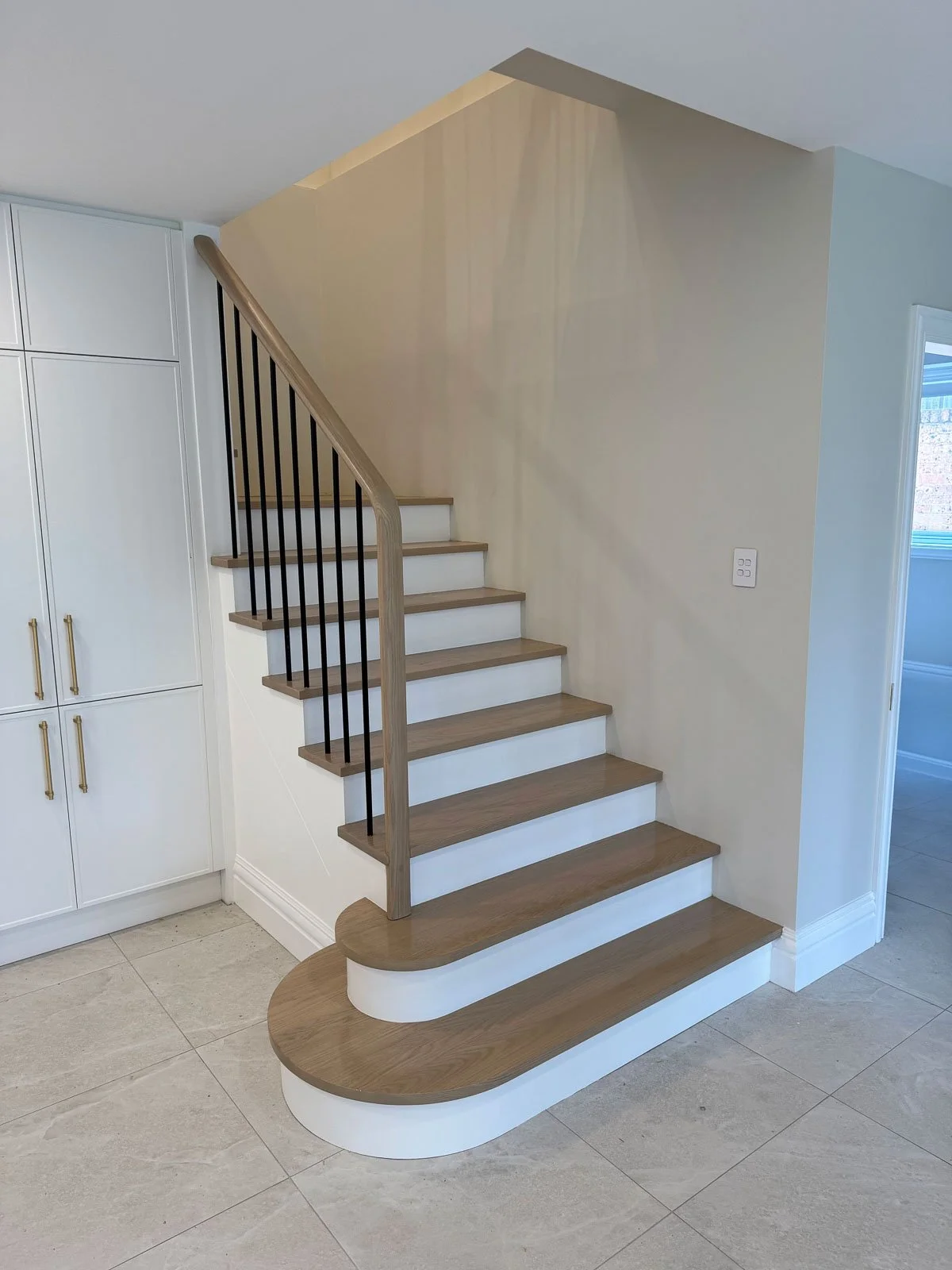 Interior view of a staircase with wooden steps, a rounded wooden base, and black metal railings, adjacent to white cabinetry and light-colored tiled flooring.