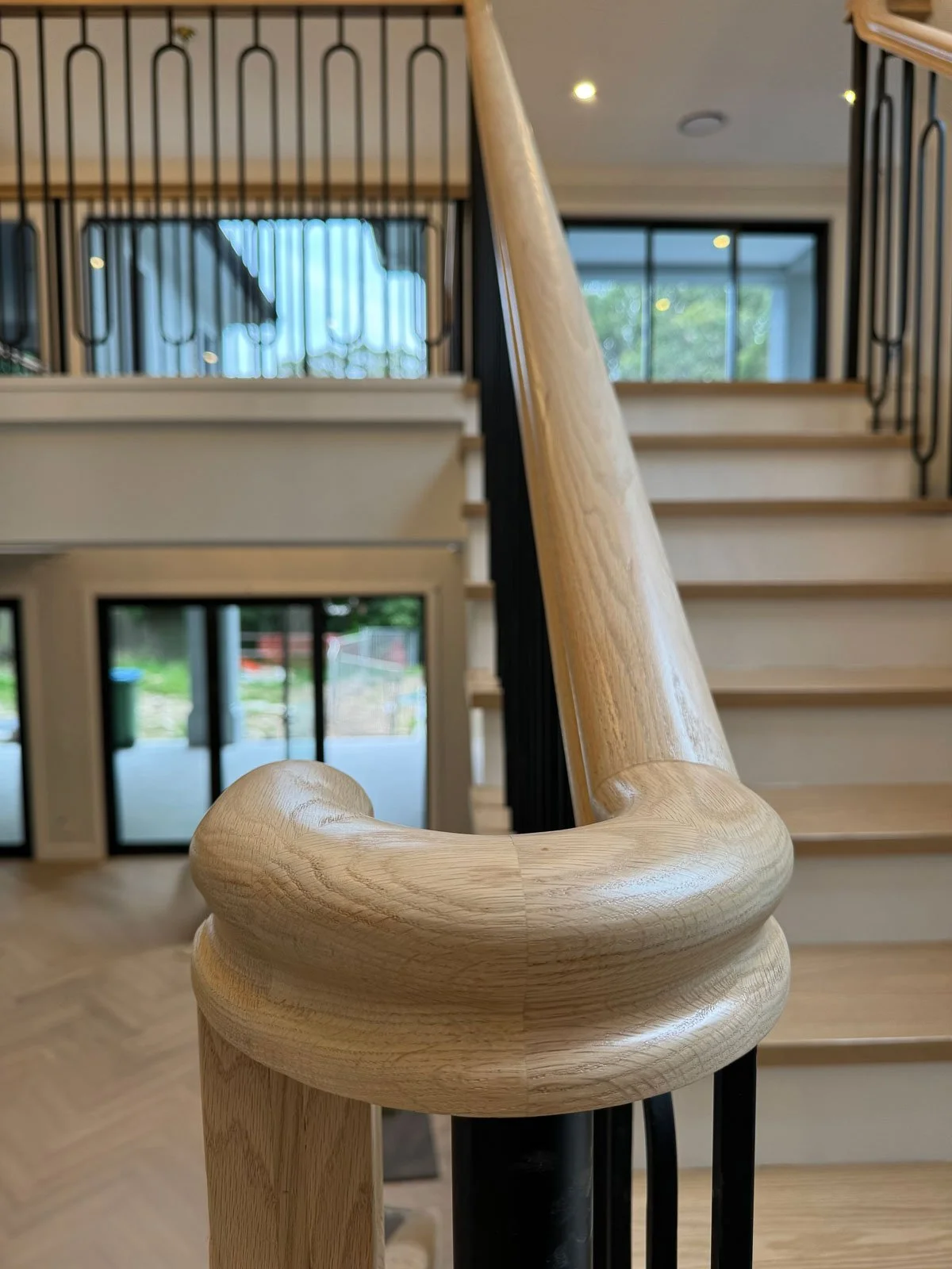 Close-up of a wooden staircase handrail in a modern home interior, with large windows and a staircase leading to the upper floor. Staircase Polishing Sydney