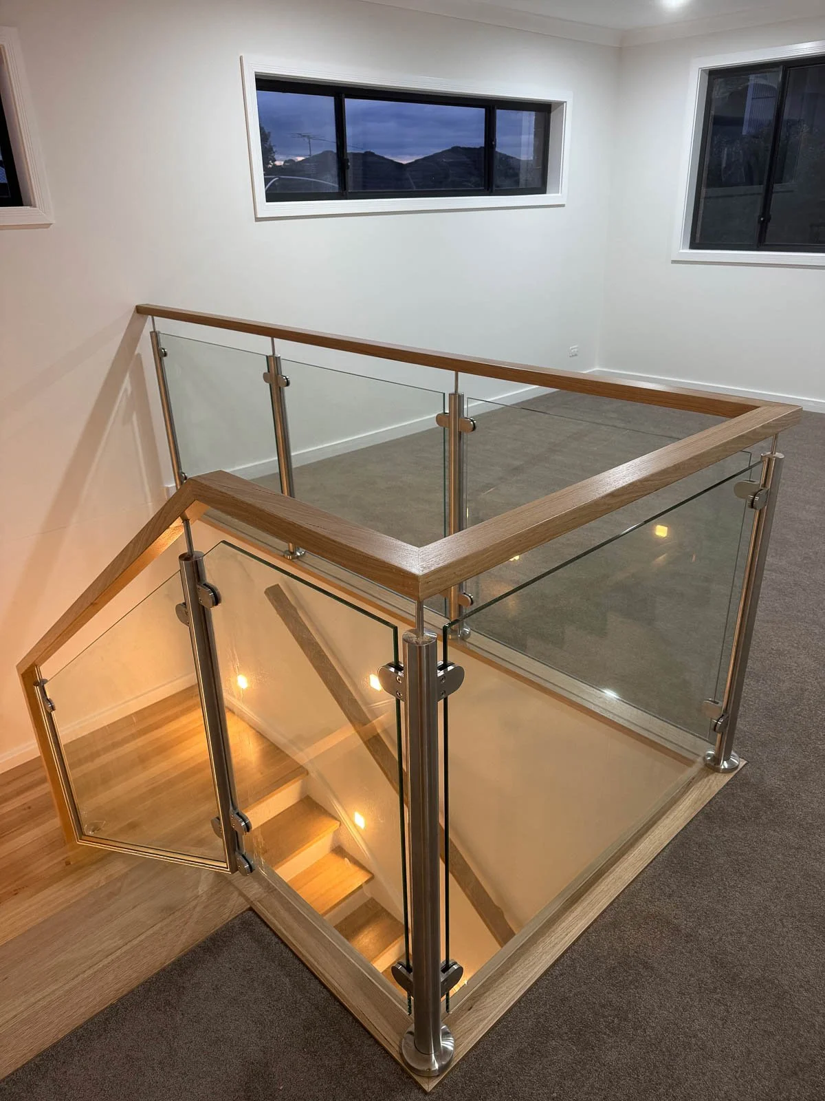 Indoor stairway with glass and wood railing, leading downstairs with illuminated steps, and overlooking a room with windows revealing an evening view of mountains. Staircase Polishing Sydney