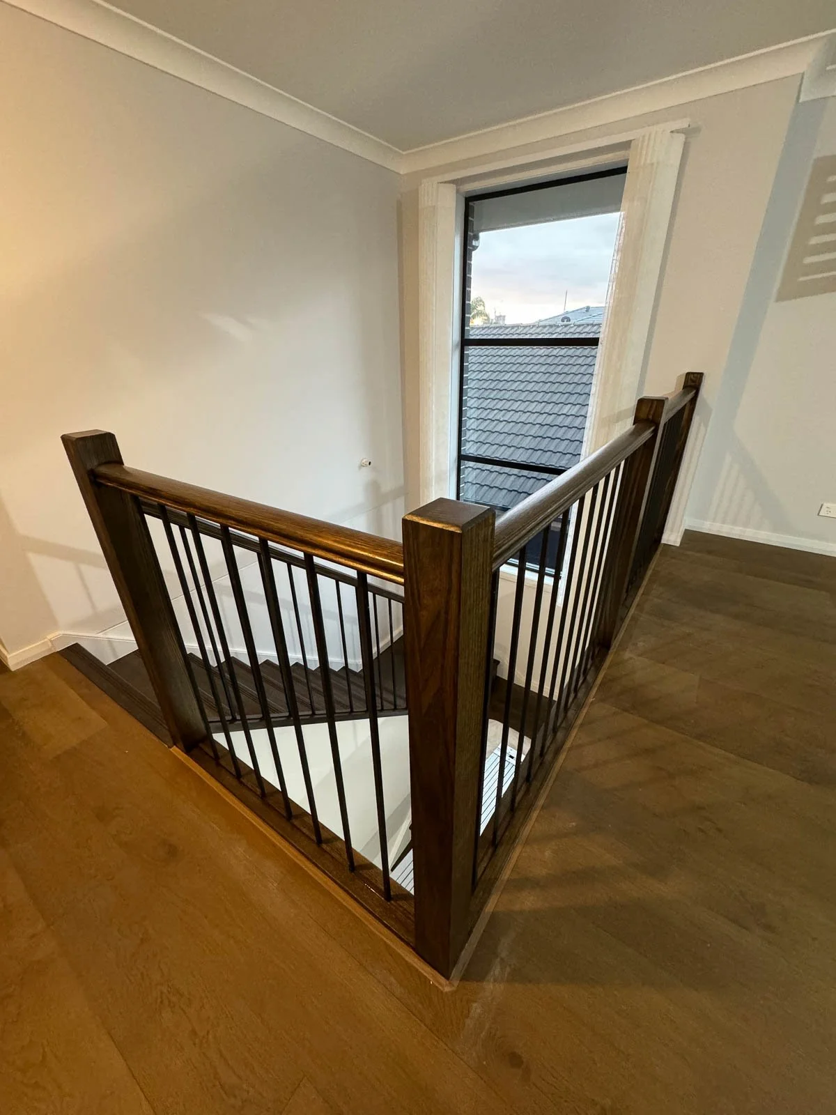 Image of an indoor staircase with wooden railing and black metal balusters, viewed from above near a window with blinds in a modern home.