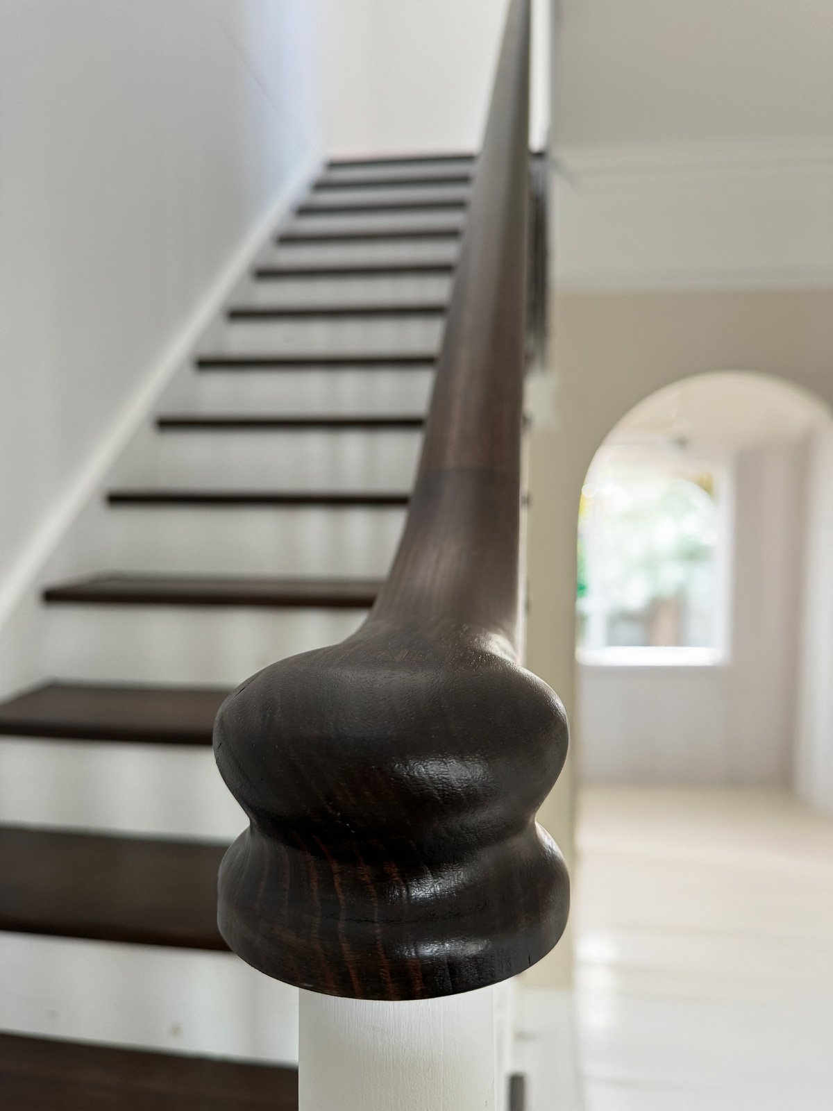 Close-up of a dark wooden stair handrail with a rounded decorative end, attached to white staircase post, with blurred background of stairs and a hallway with a door and window. Staircase Polishing Sydney