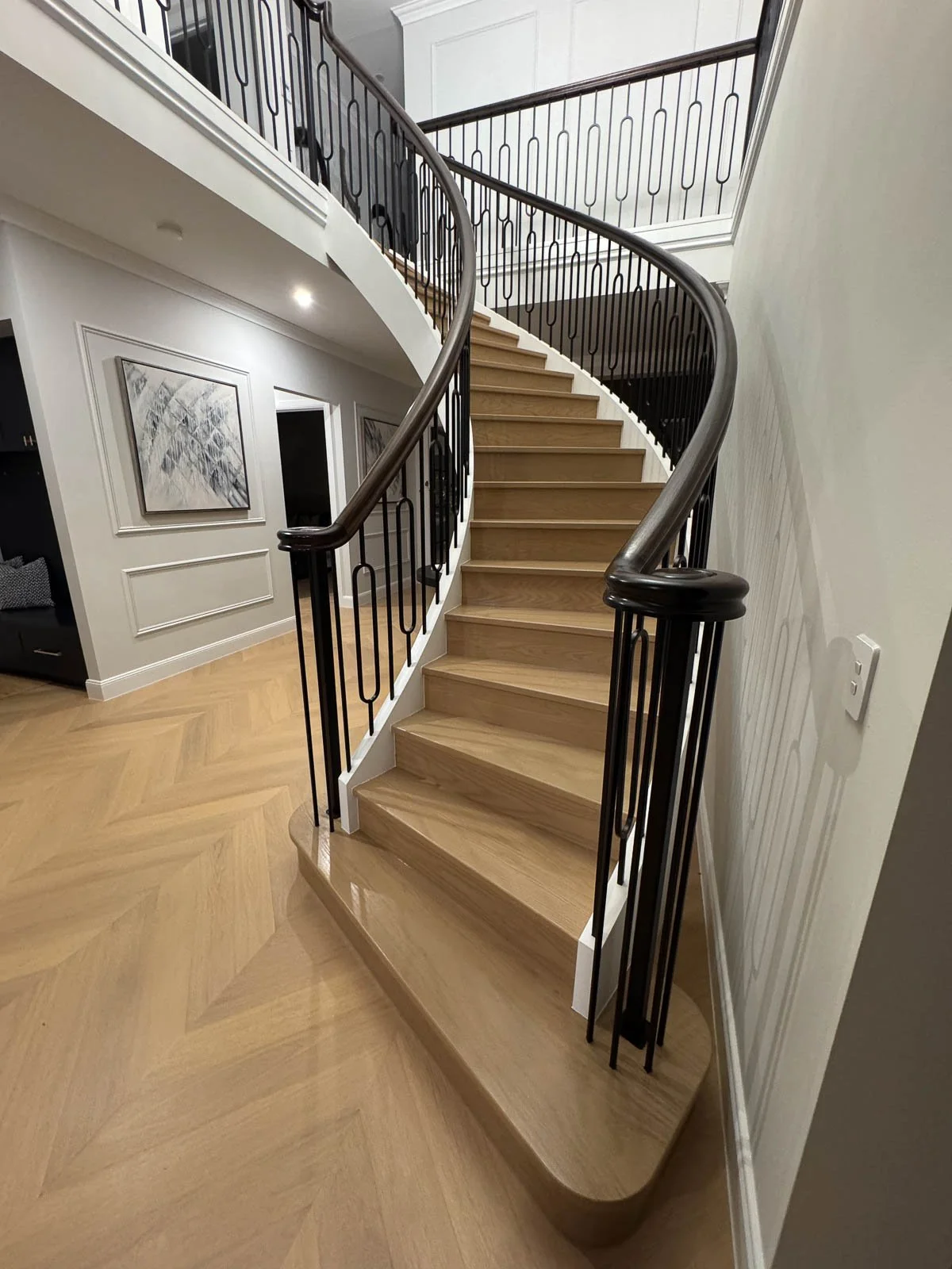 Interior view of a curved staircase with wooden steps, black metal railings, and a polished wooden handrail in a modern home. Staircase Polishing Sydney