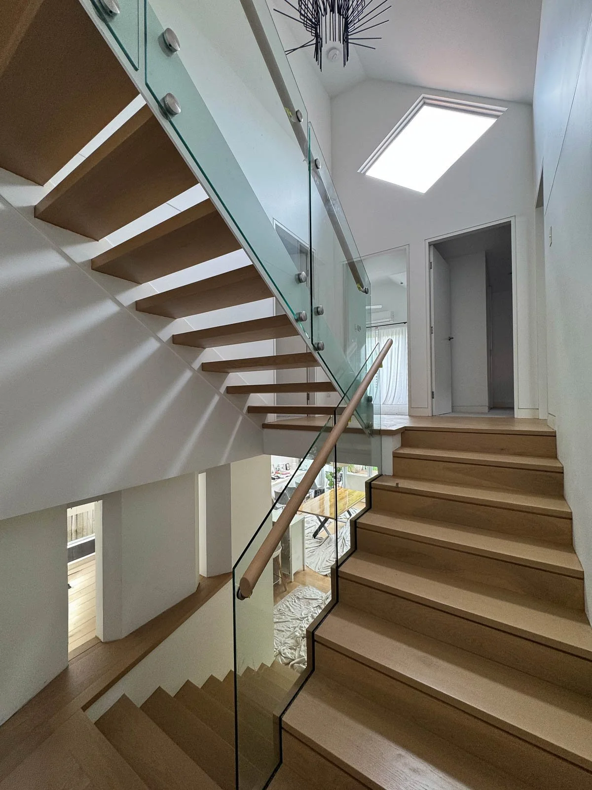 Modern staircase with wooden steps and glass railing inside a house, with skylight and decorative ceiling light. Staircase Polishing Sydney