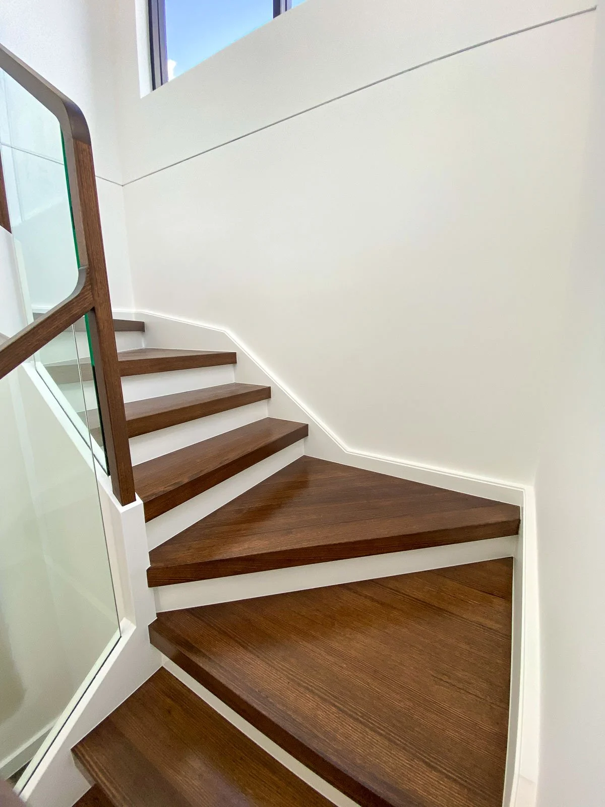 Photo of a modern wood staircase with dark wood steps and a glass railing, visible from the lower to the upper level, with white walls and a small window at the top letting in natural light. Staircase Polishing Sydney