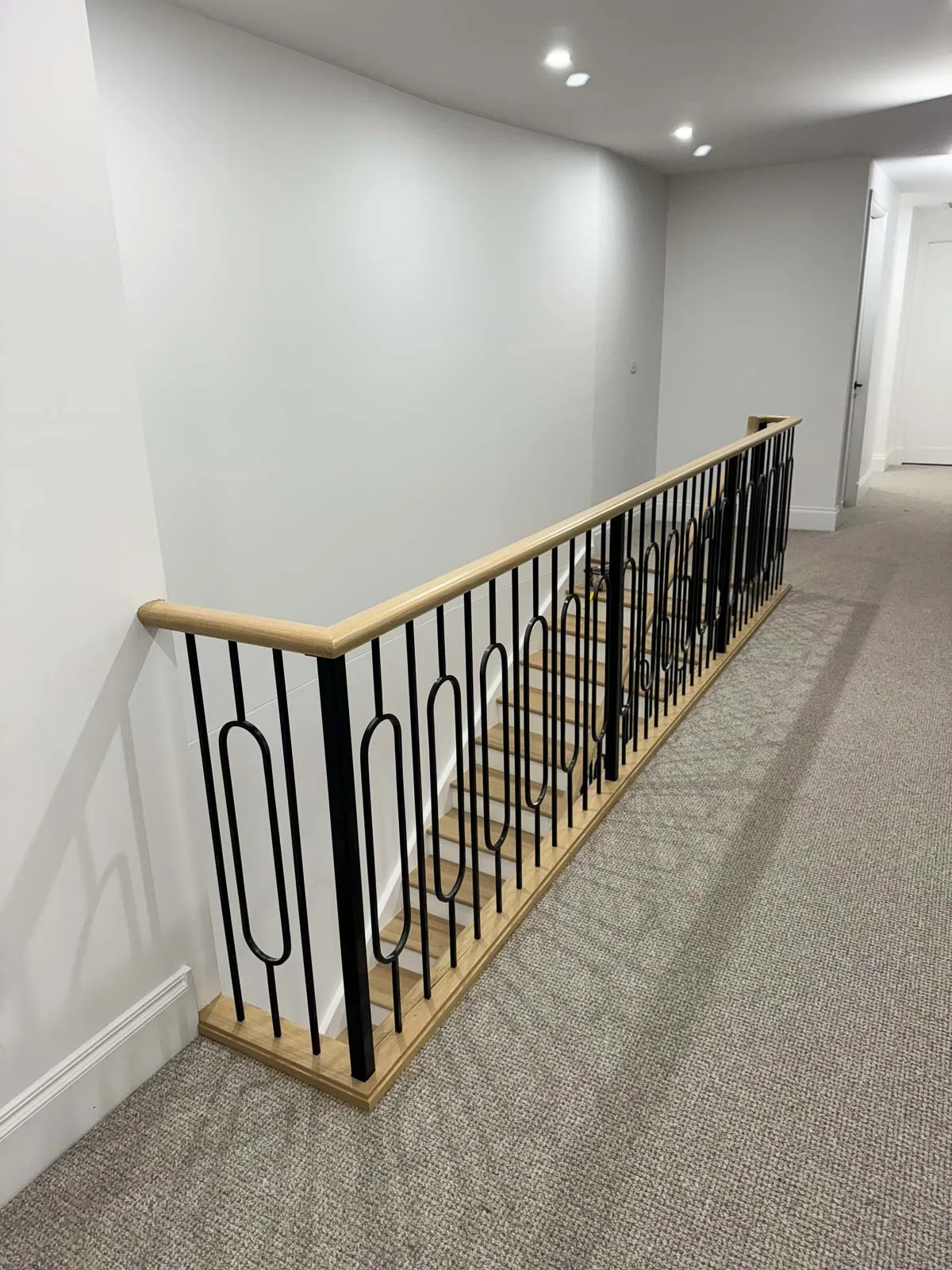 Indoor staircase with black metal railing and wooden handrail, leading down to a lower level, with white walls and grey carpeted floor. Staircase Polishing Sydney