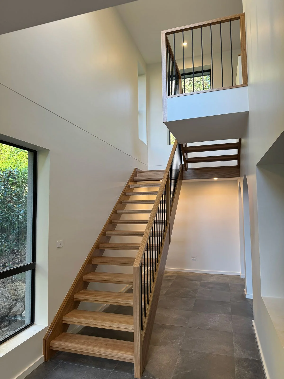 Interior view of a modern house featuring a wooden staircase with black metal railing, large window to the left allowing natural light, and a second-floor balcony with matching railing. Staircase Polishing Sydney