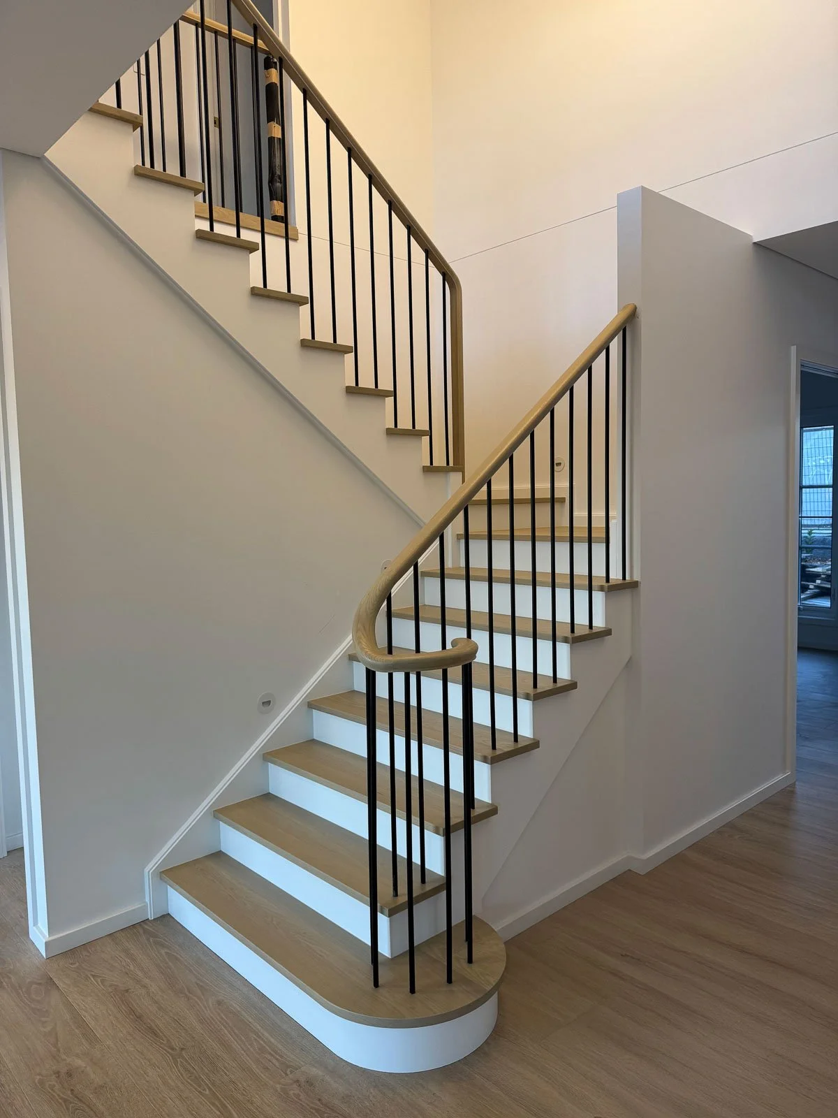 Interior staircase with light wood treads, black metal balusters, and a curved wooden handrail, leading to an upper floor in a modern home. Staircase Polishing Sydney