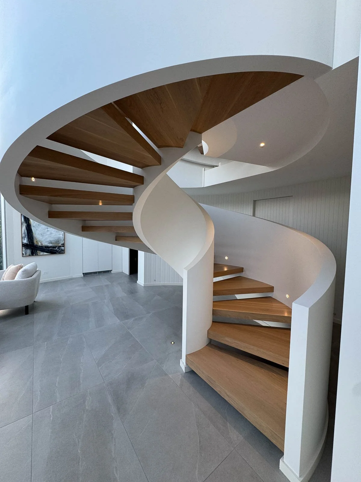 Interior view of a modern, curved staircase with wooden steps and white side panels, leading upwards inside a contemporary home with gray tiled floors and minimalistic decor. Staircase Polishing Sydney