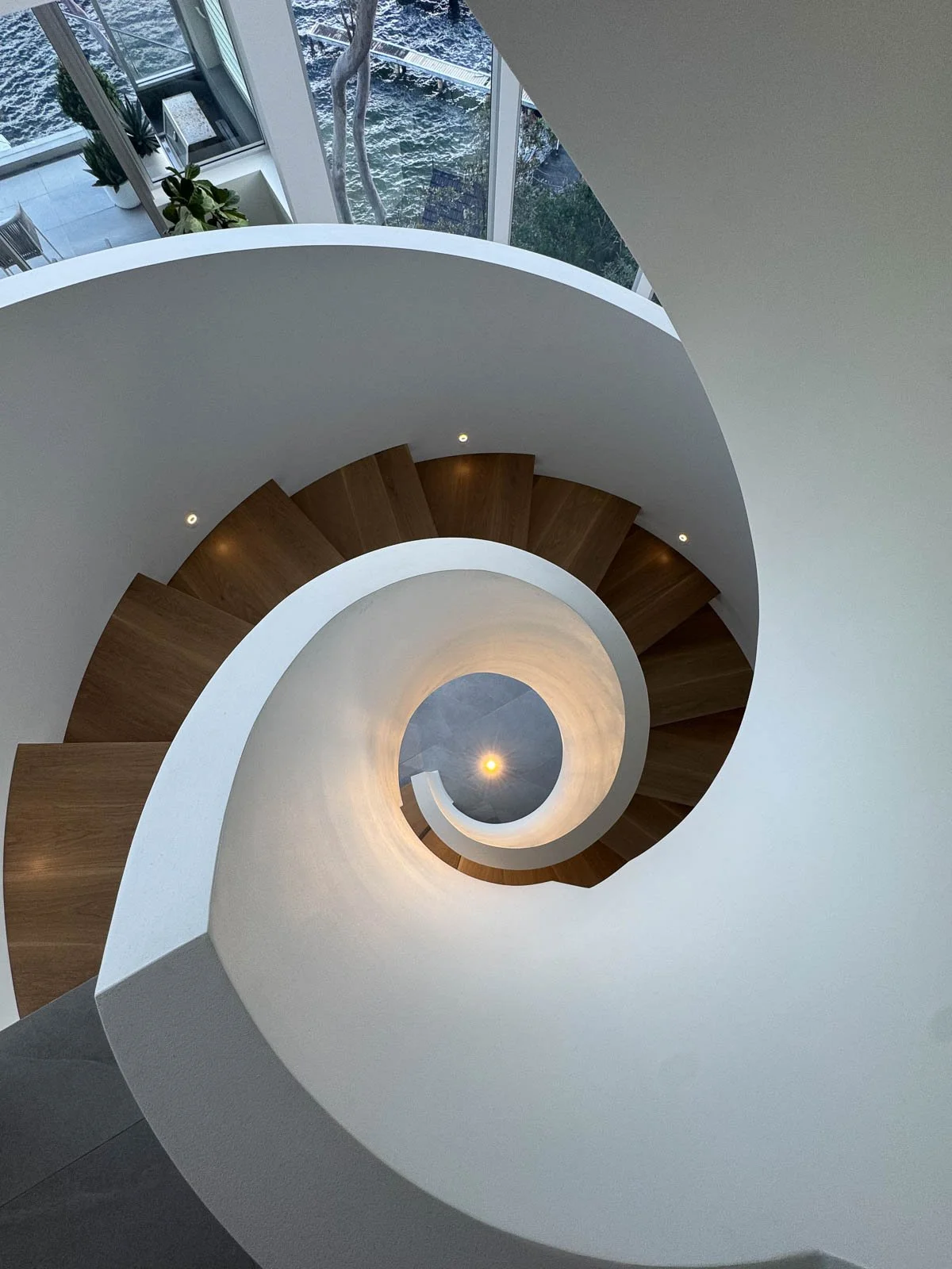 Top-down view of a spiral staircase with wooden steps and white walls, with a window showing a river and trees outside. Staircase Polishing Sydney