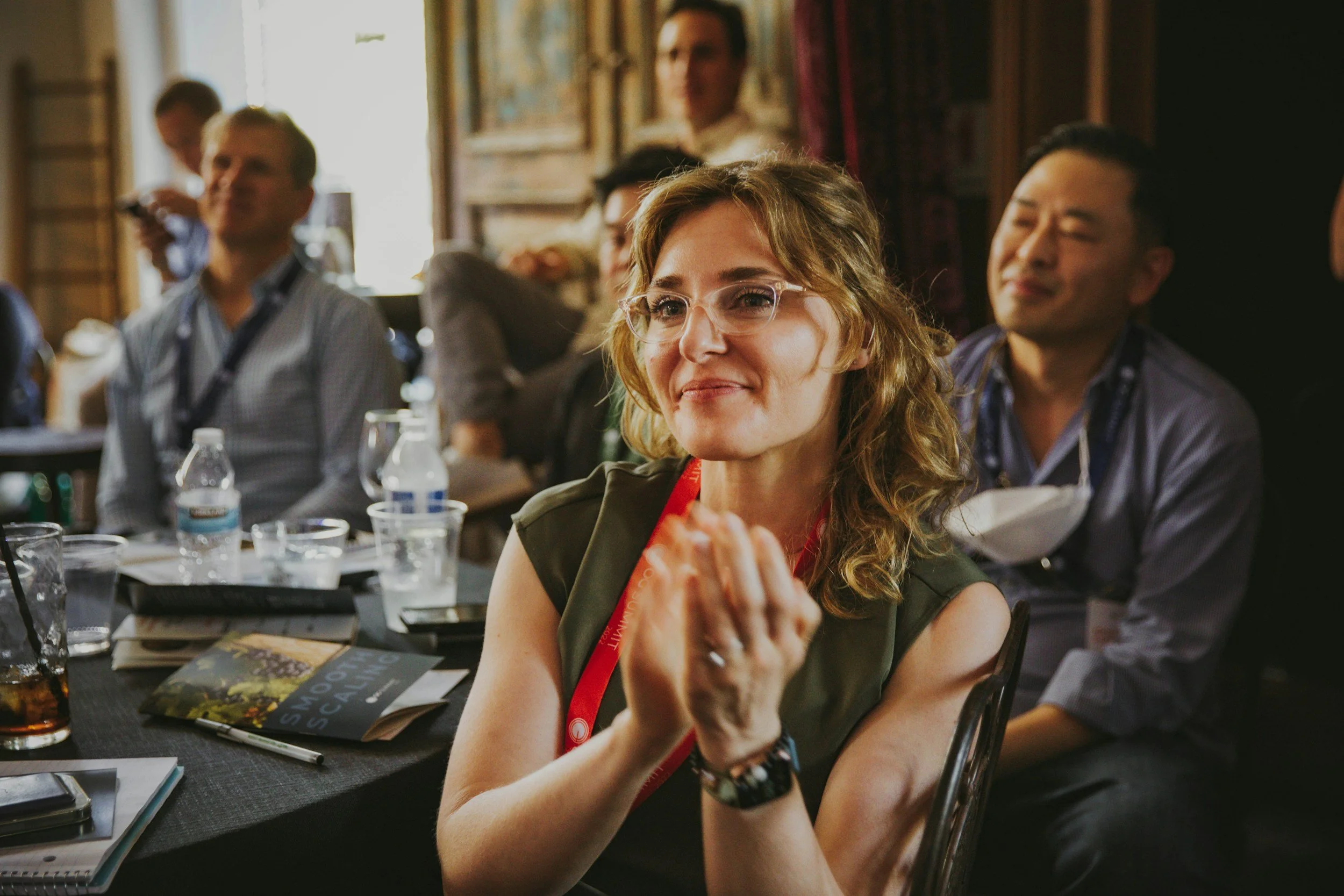 A group of people attending a conference or seminar, sitting at a long table with notebooks, books, and water bottles, clapping and smiling. The woman in the foreground has glasses, curly hair, and wears a watch, while others are blurred in the background.