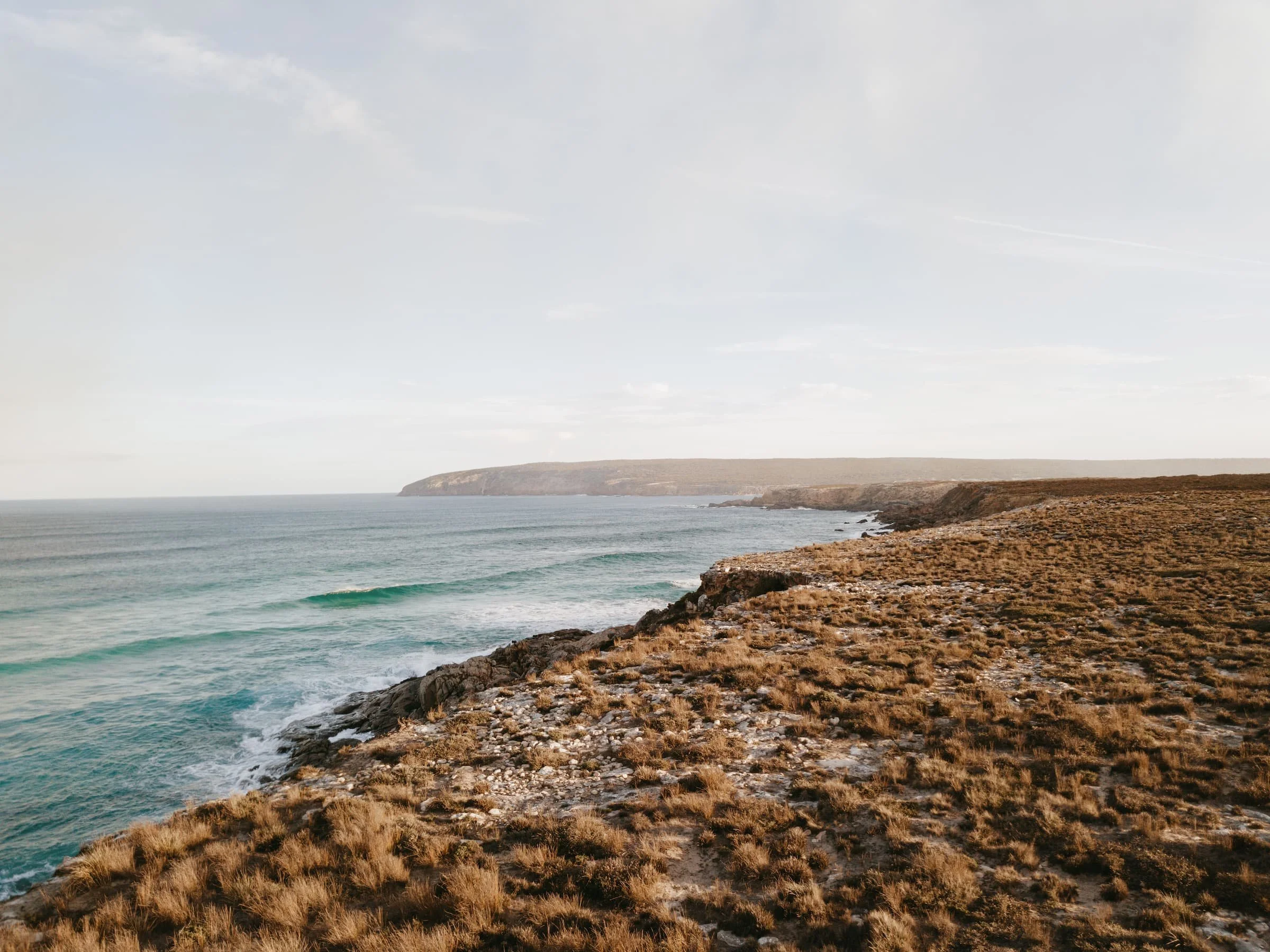 Coastal scene with rocky shore, sparse dry grass, calm ocean with small waves, and a distant headland under a partly cloudy sky.