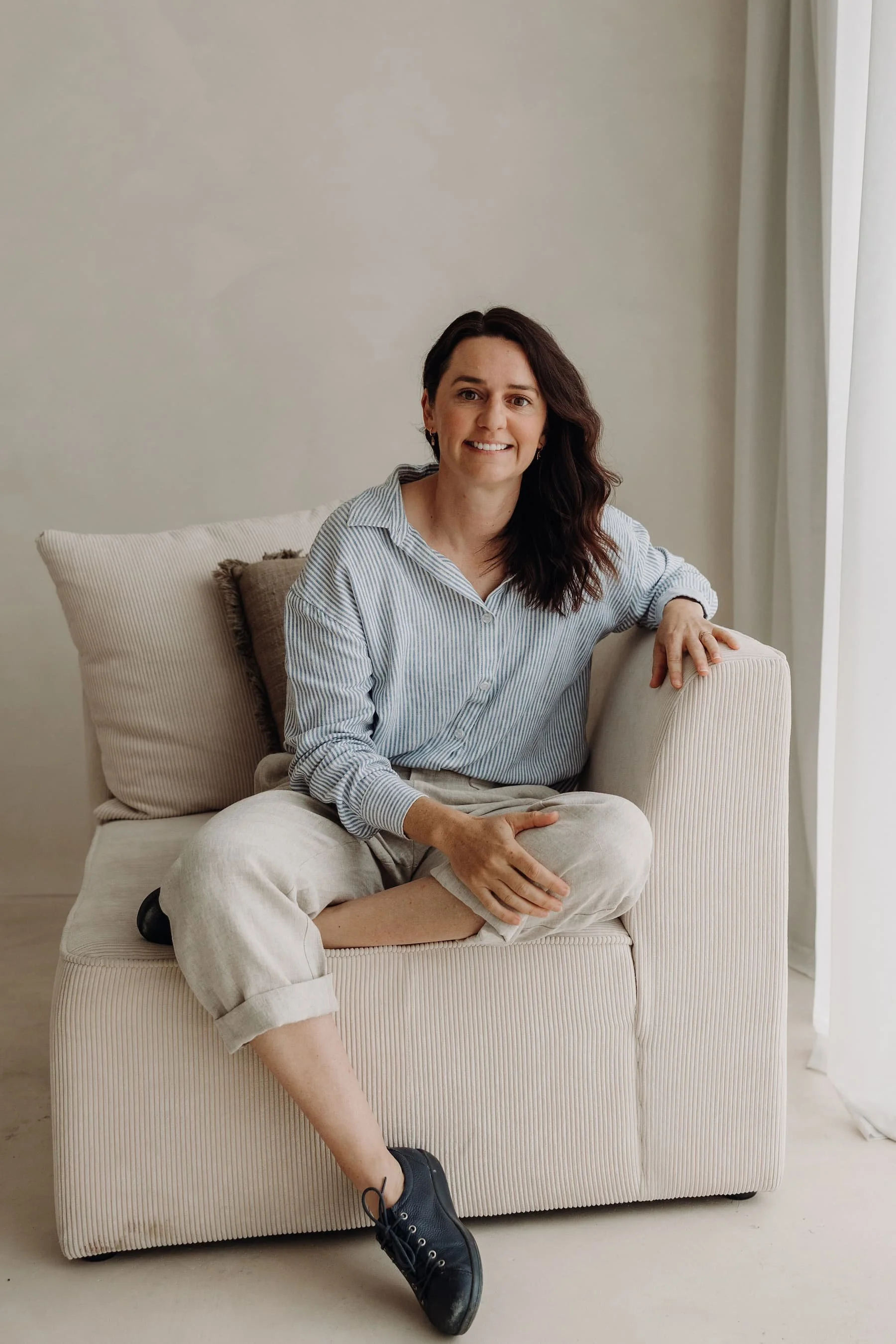 A woman with dark wavy hair, wearing a light striped shirt and beige pants, is sitting on a cream-colored sofa, smiling at the camera.