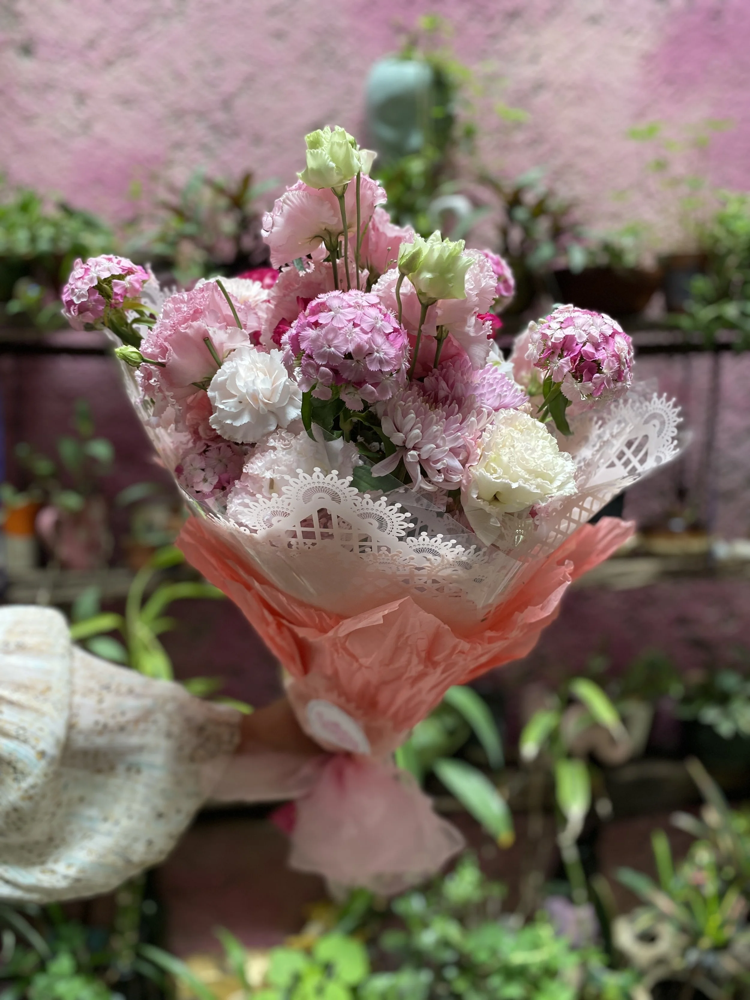 A hand holding a pink bouquet of mixed flowers wrapped in pink tissue paper, with a blurred garden background.
