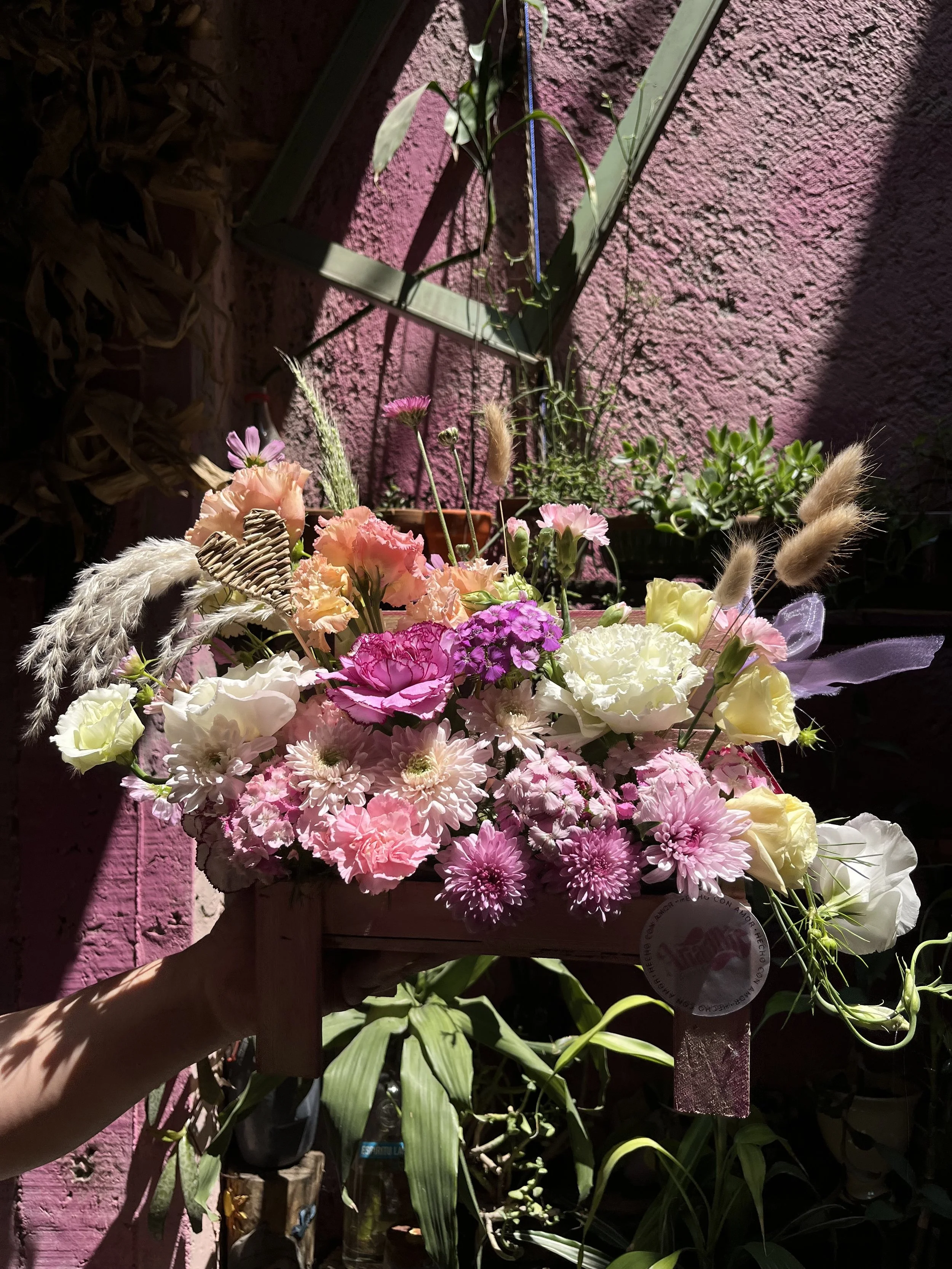 A colorful flower arrangement in a wooden box held by a hand, with pink, white, and purple flowers, dried grasses, and a decorative butterfly, set against a pink textured wall with sunlight and shadows.