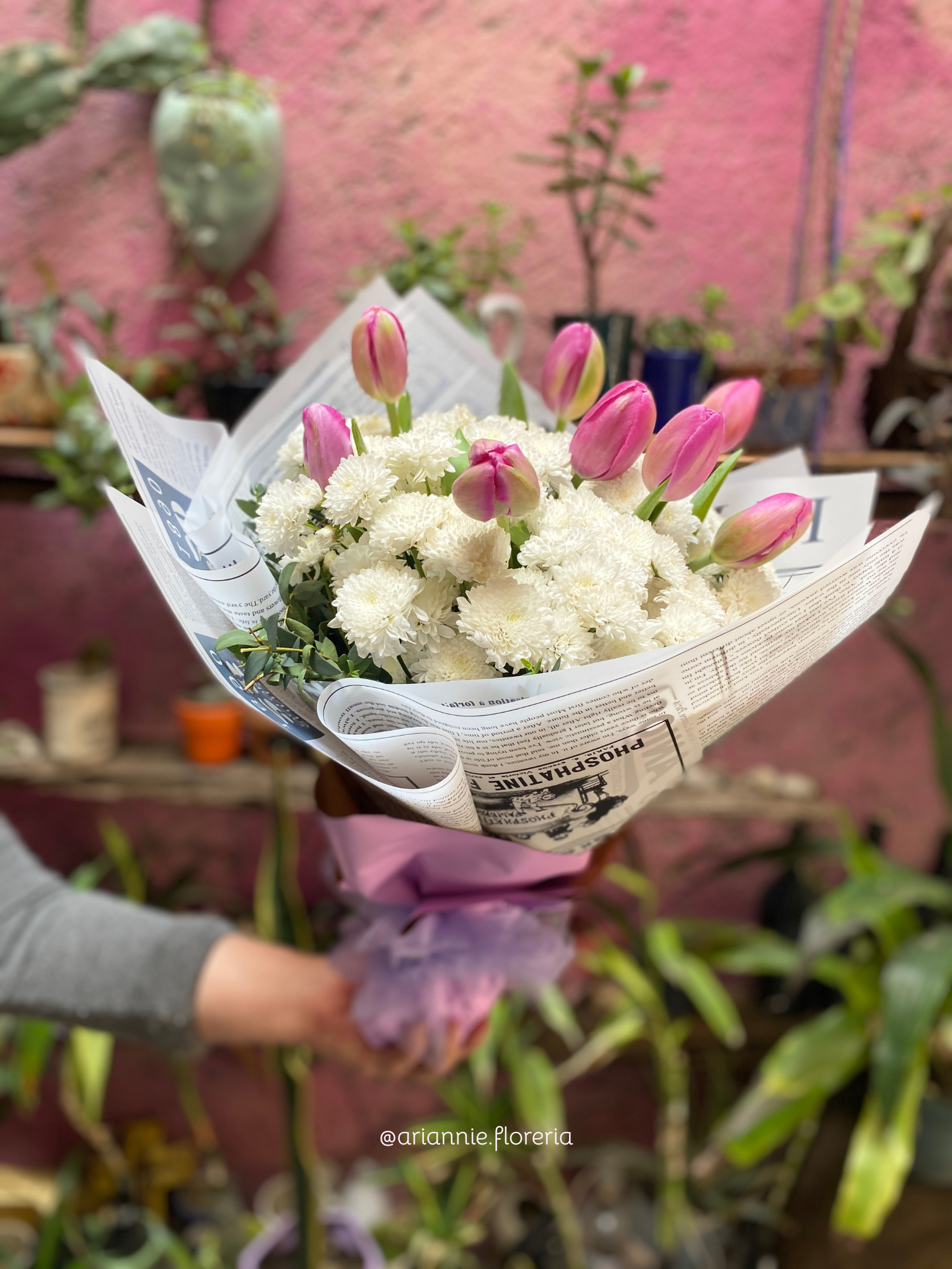 A person holding a bouquet of pink tulips and white chrysanthemums wrapped in newspaper and pink paper, with a garden background.