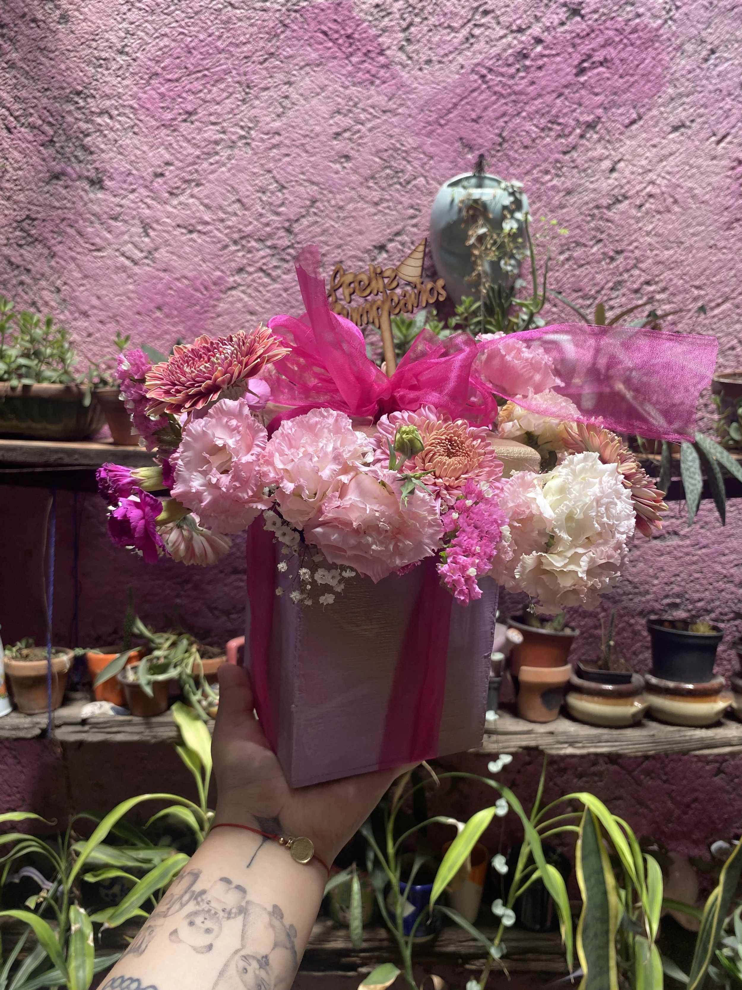 A hand holding a pink gift box decorated with flowers and pink ribbons, with a pink textured wall in the background.