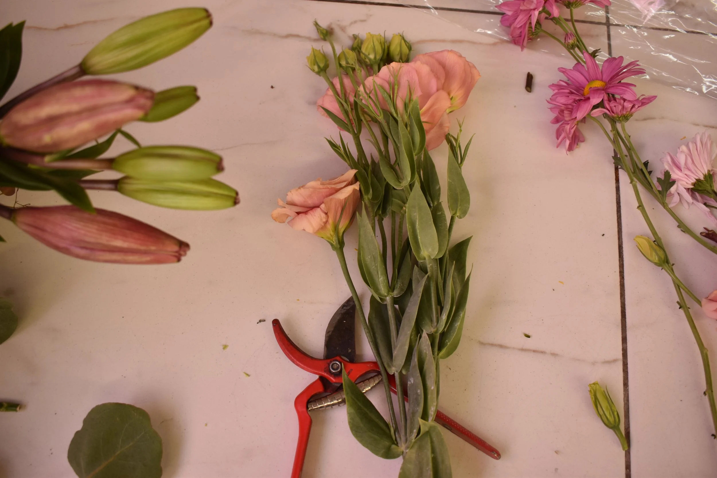 Flowers in process of arranging on table, including pink lilies, pink daisies, and peach roses, with floral pruning tools nearby.