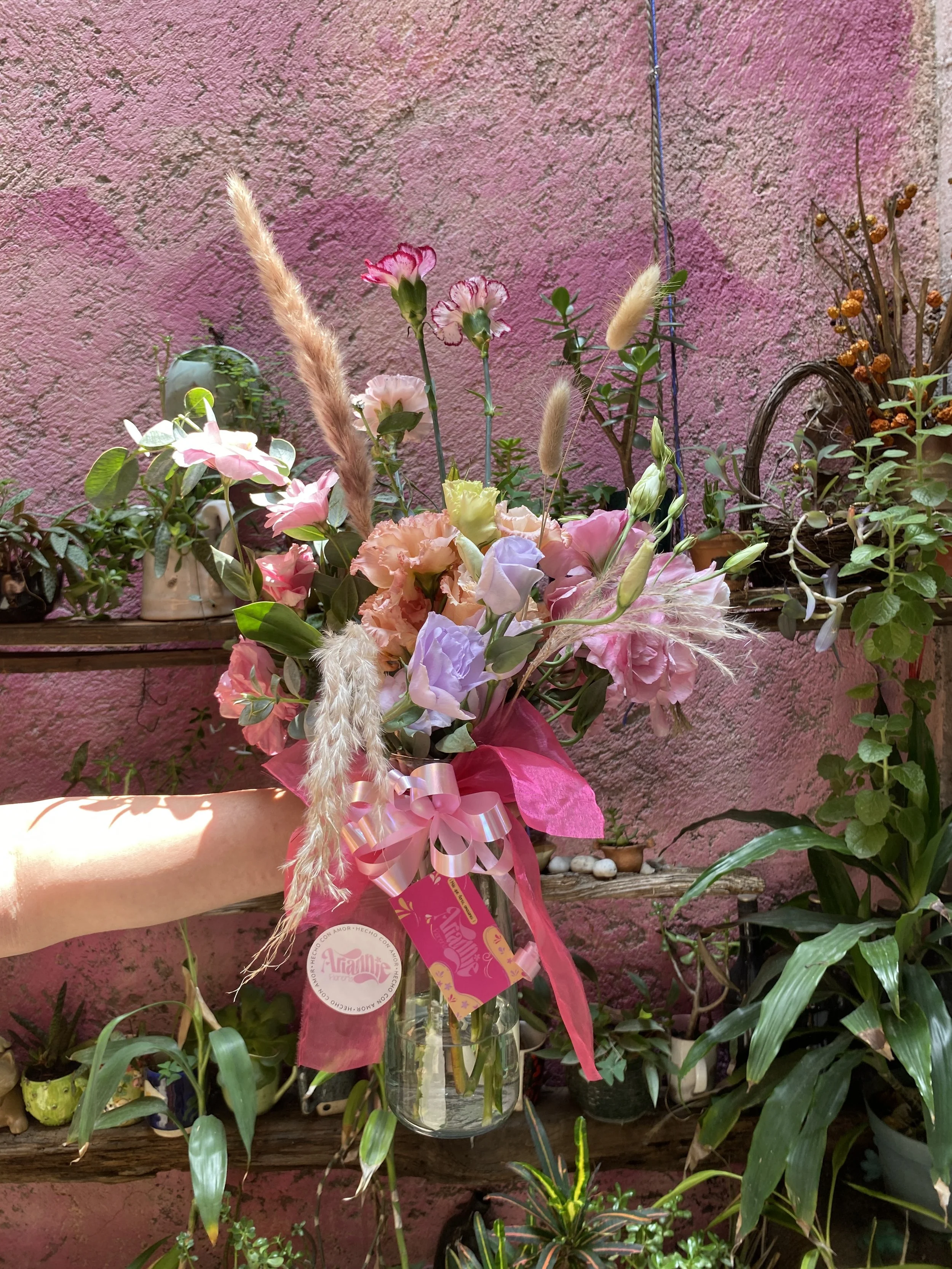 A colorful flower bouquet with pink, white, purple, and peach roses, various grasses, and greenery, wrapped with pink ribbon, held in front of a pink textured wall with indoor plants and shelves.