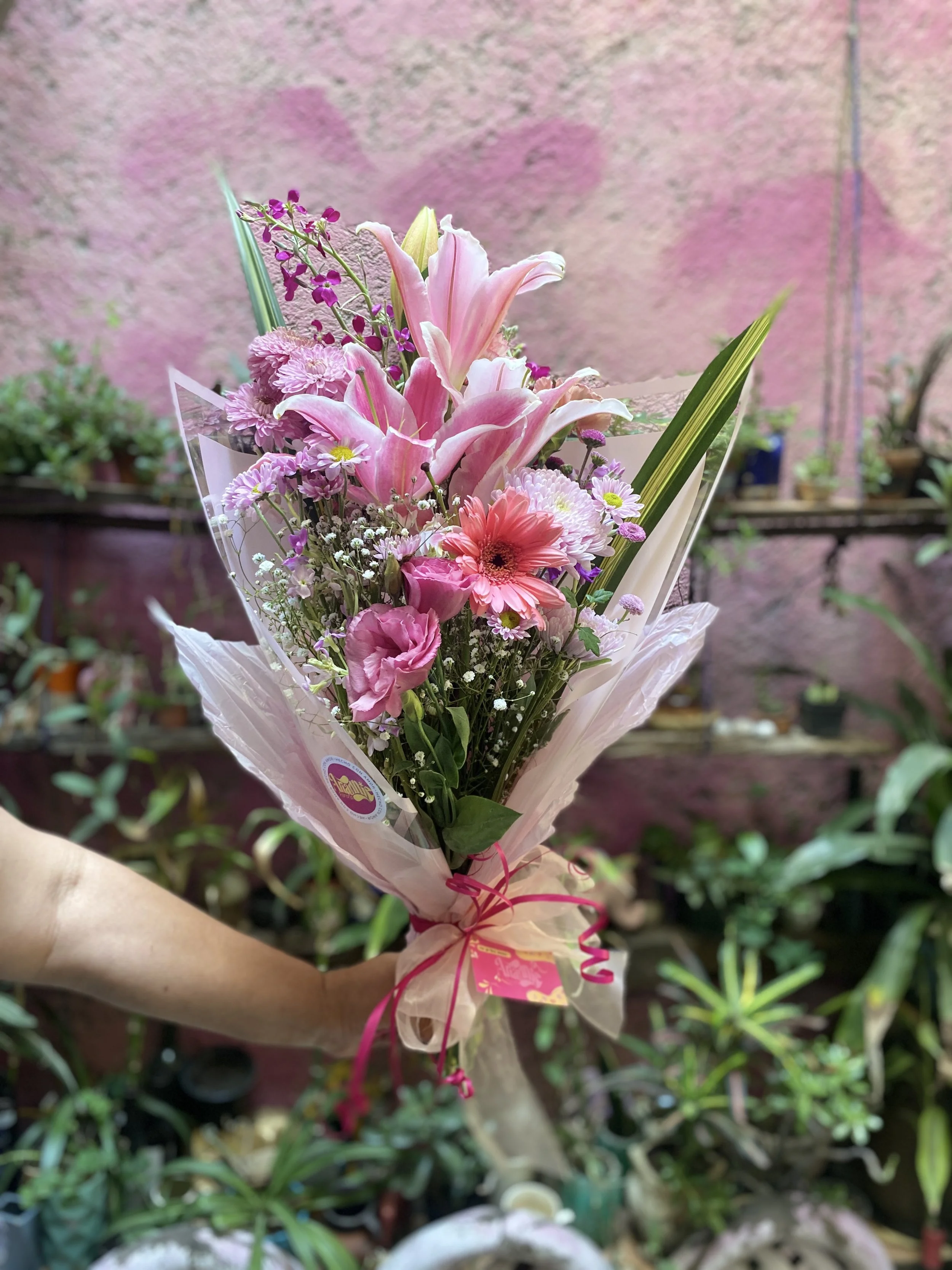 A hand holding a bouquet of pink lilies, gerbera daisies, and other mixed flowers wrapped in pink and clear wrapping paper.