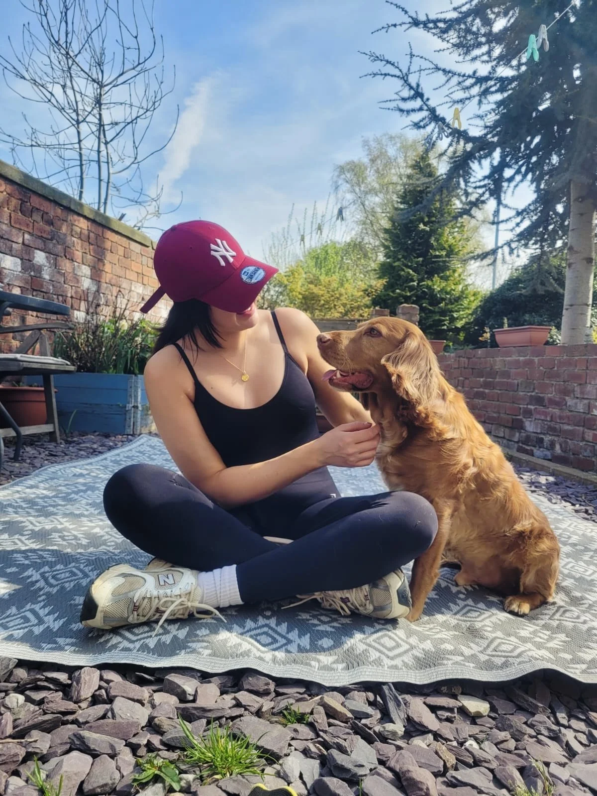 A woman in black athletic clothing, sitting cross-legged on a patterned outdoor blanket, is interacting with a golden retriever dog, in a backyard with brick walls, potted plants, and trees under a blue sky.