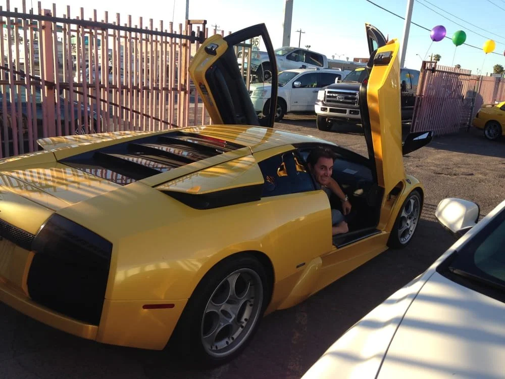 A yellow sports car with its gullwing doors open, parked in a lot with other cars, and a person sitting inside smiling.