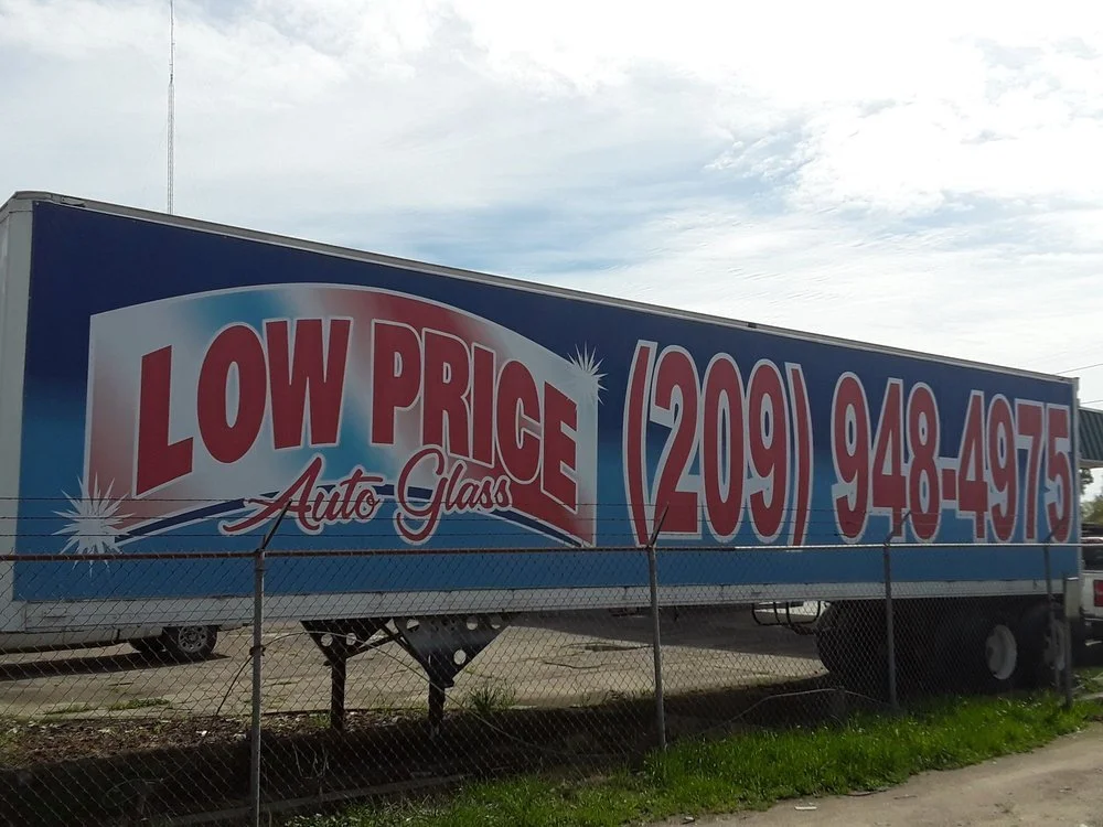 Large advertisement billboard for auto glass with the text 'LOW PRICE Auto Glass' and a phone number (209) 948-4975. The billboard is blue with red and white lettering, behind a chain-link fence, under a partly cloudy sky.