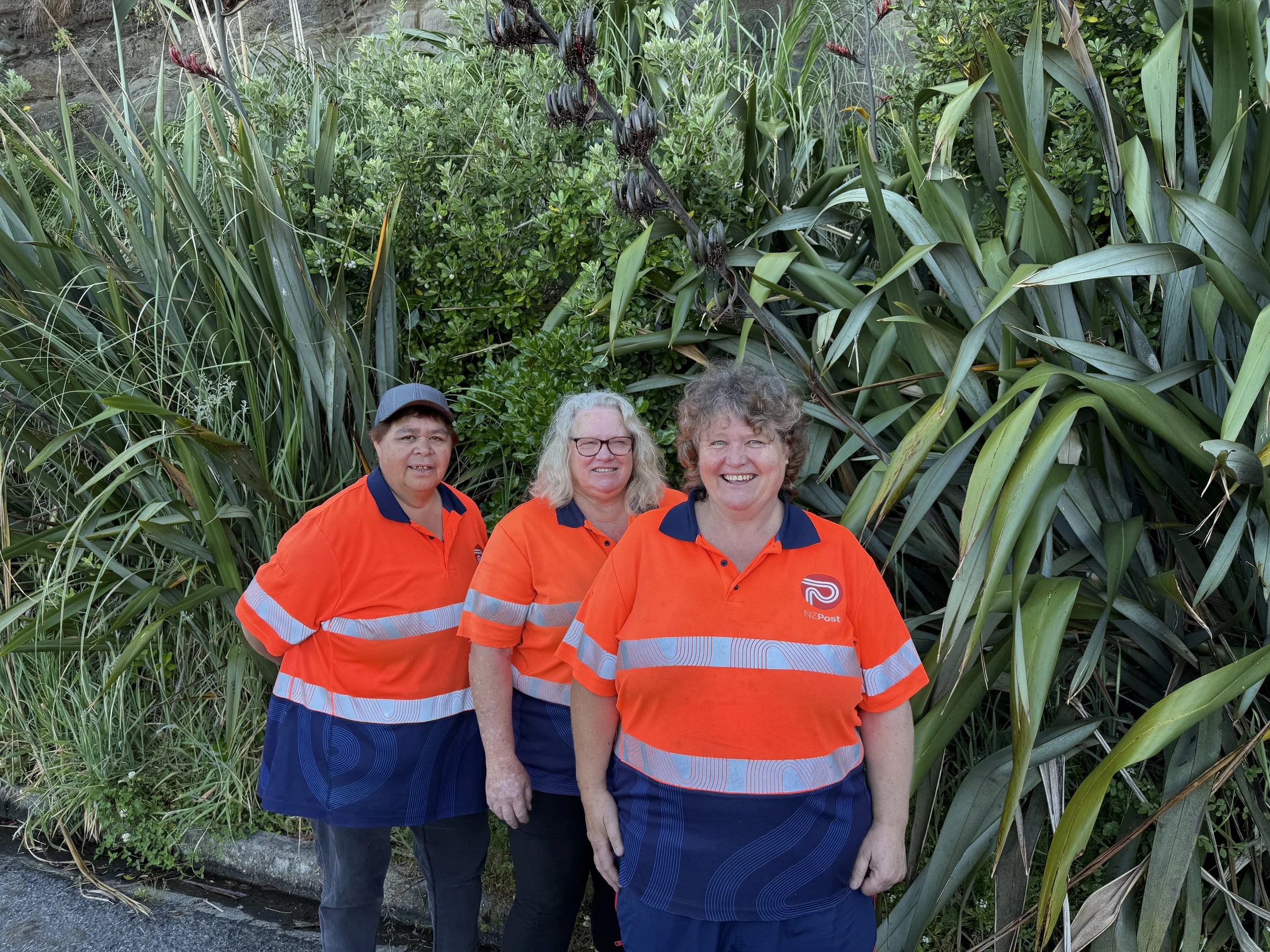 Three NZ Post employees in orange uniforms standing in front of lush green plants and foliage, smiling for the camera.