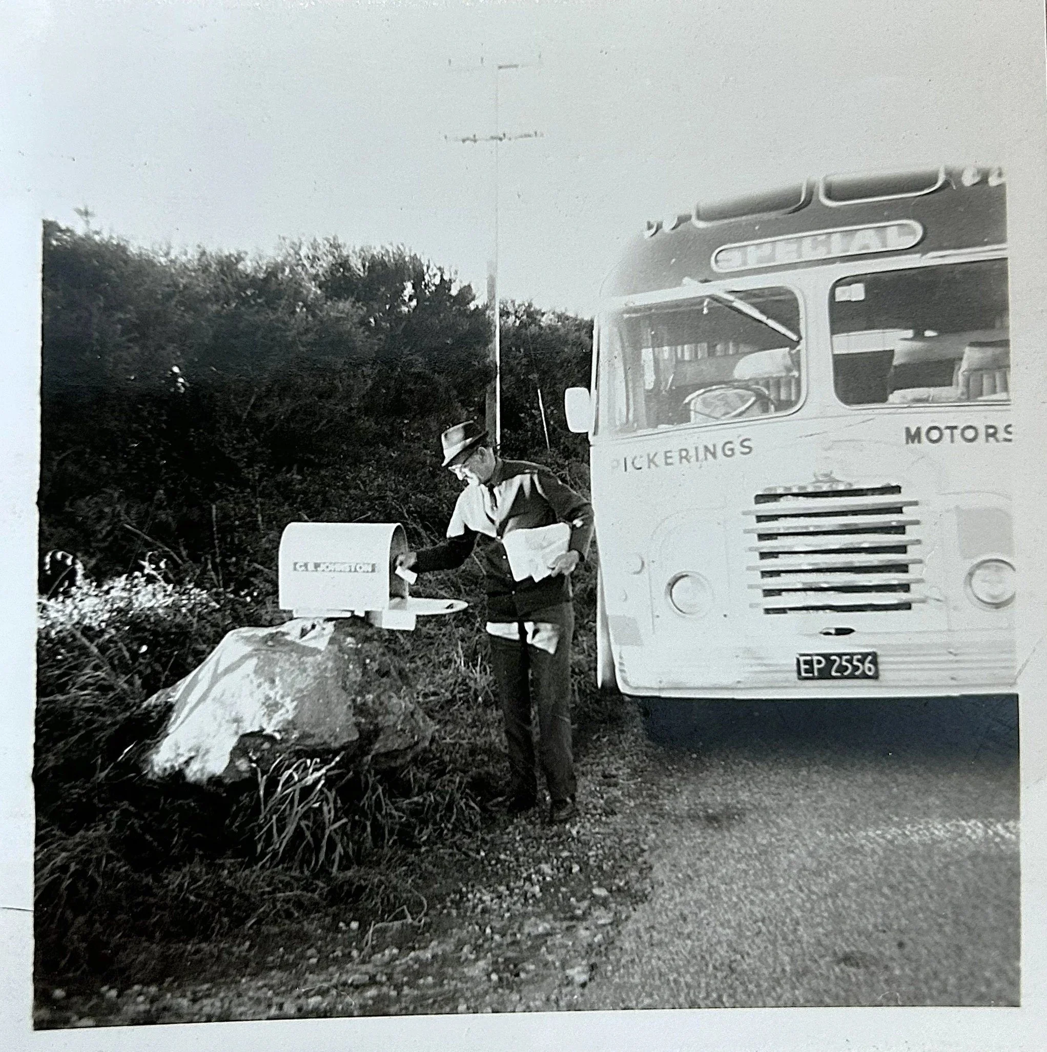 A person dressed in vintage clothing, wearing glasses and a hat, is placing a letter into a mailbox on a large rock. A large bus with the sign 'Clickings Motors' is parked nearby.