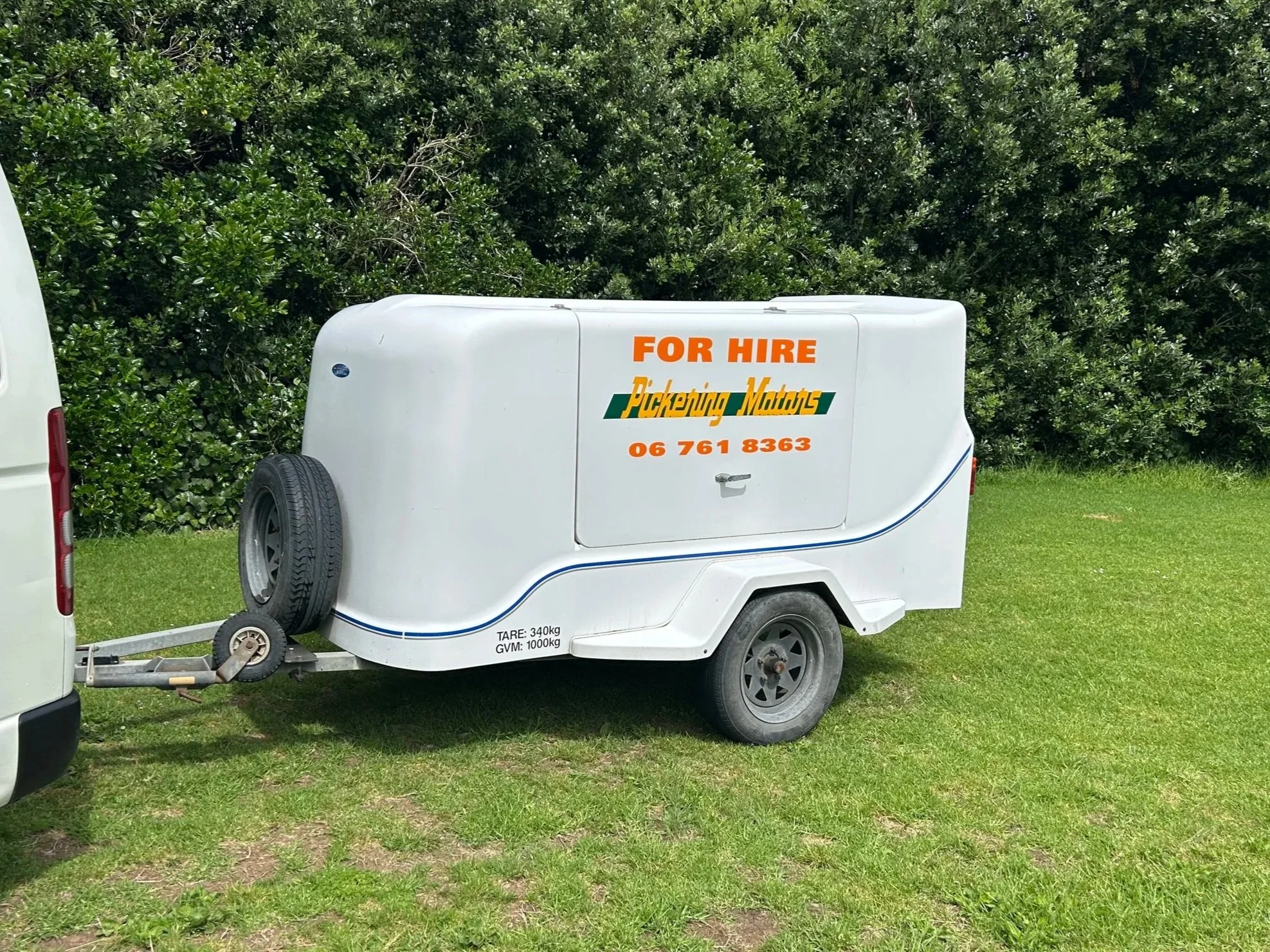 Small white horse trailer with orange and green lettering that says 'For Hire Pickering Motors' and a phone number, attached to a vehicle on a grassy area with bushes and trees in the background.
