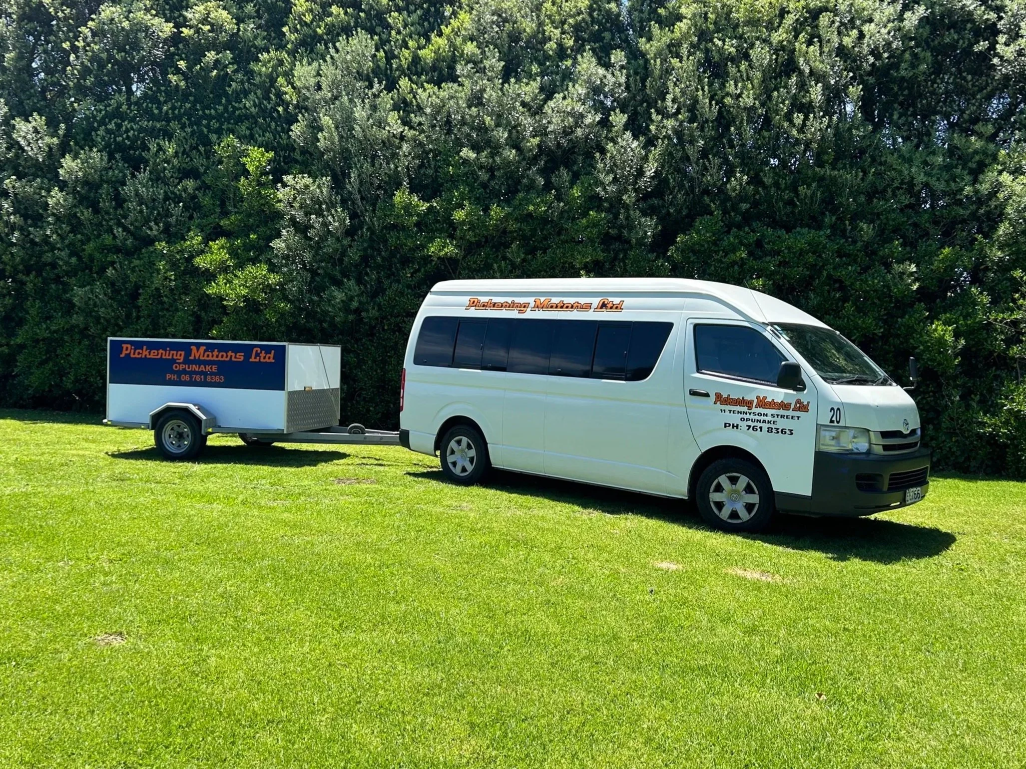 White van with company logo and contact information pulling a small trailer on grass with bushes in the background.