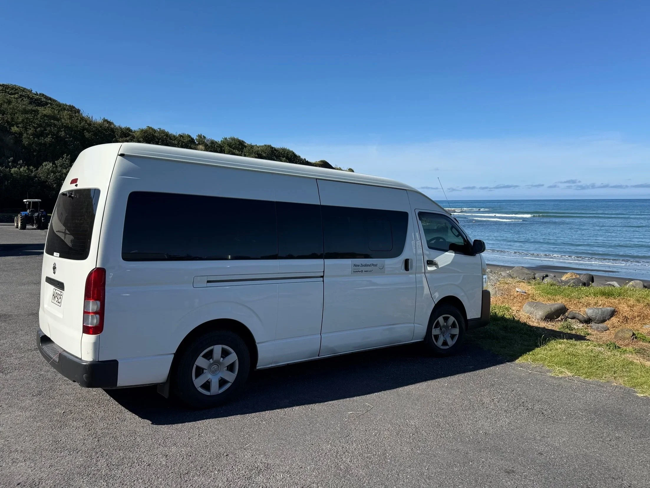 A white van parked near a beach with ocean waves, rocks, and a clear blue sky.