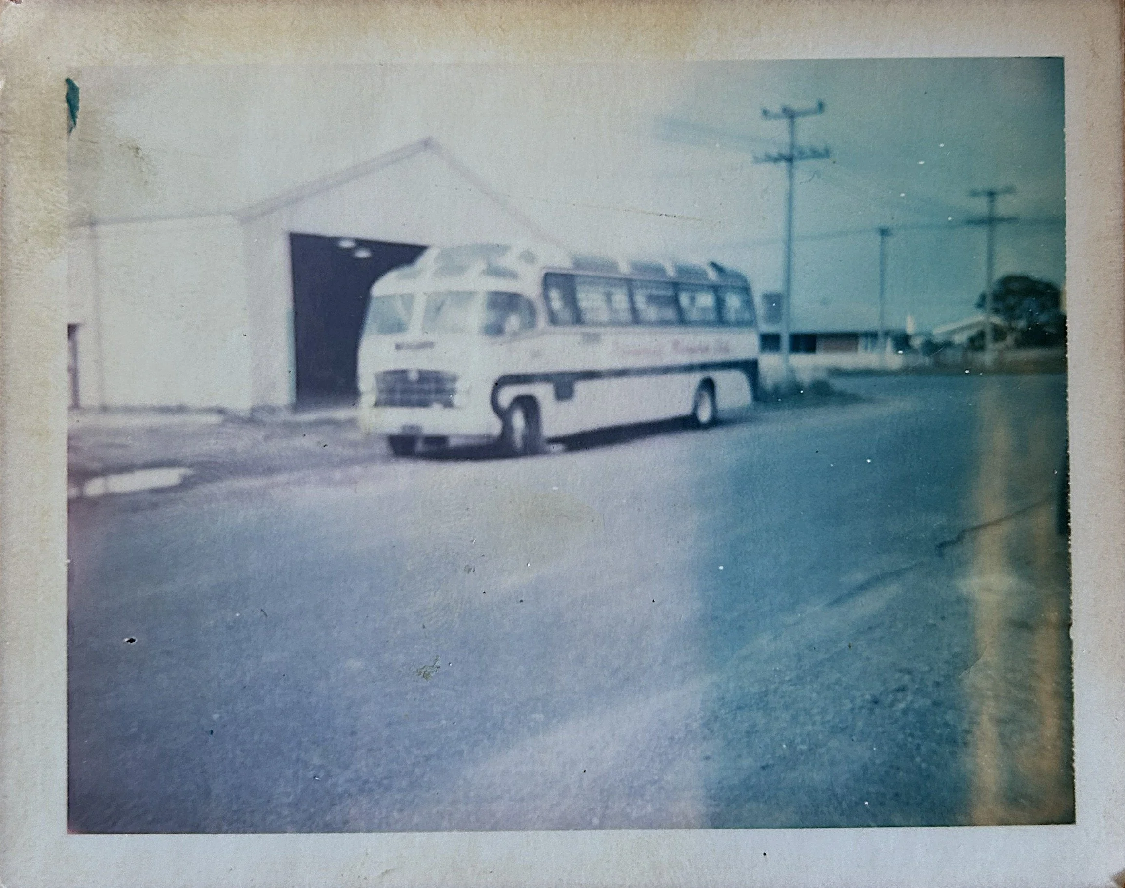 Black and white vintage photo of a bus parked in front of a garage with a wide open door, utility poles and some buildings in the background.