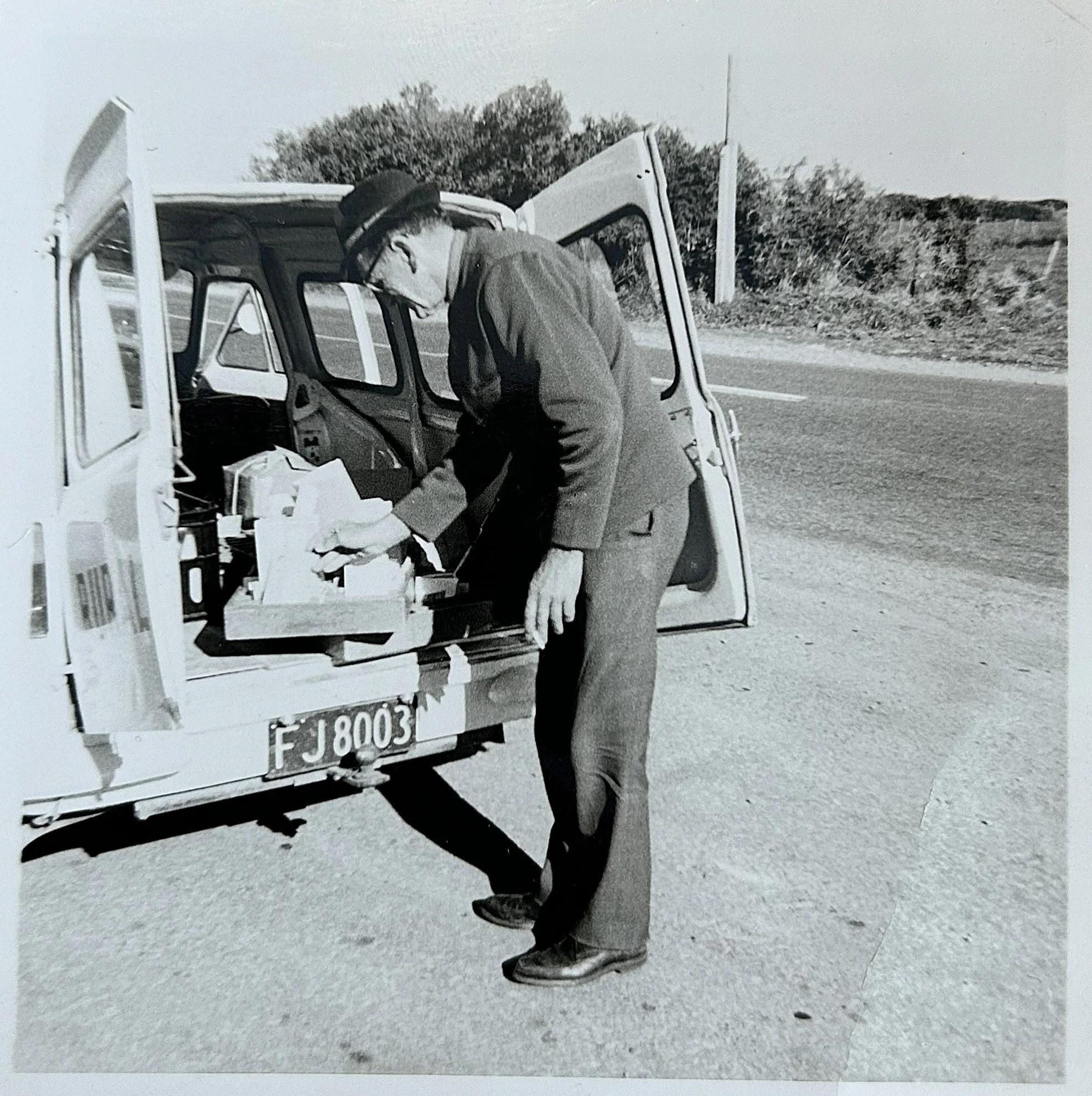 A man dressed in a suit, hat, and glasses, is leaning into the open back of a small vehicle, possibly a vintage van or truck, looking at items inside. The vehicle has a visible license plate number 'FJ 8003' and is parked on the side of a rural road 