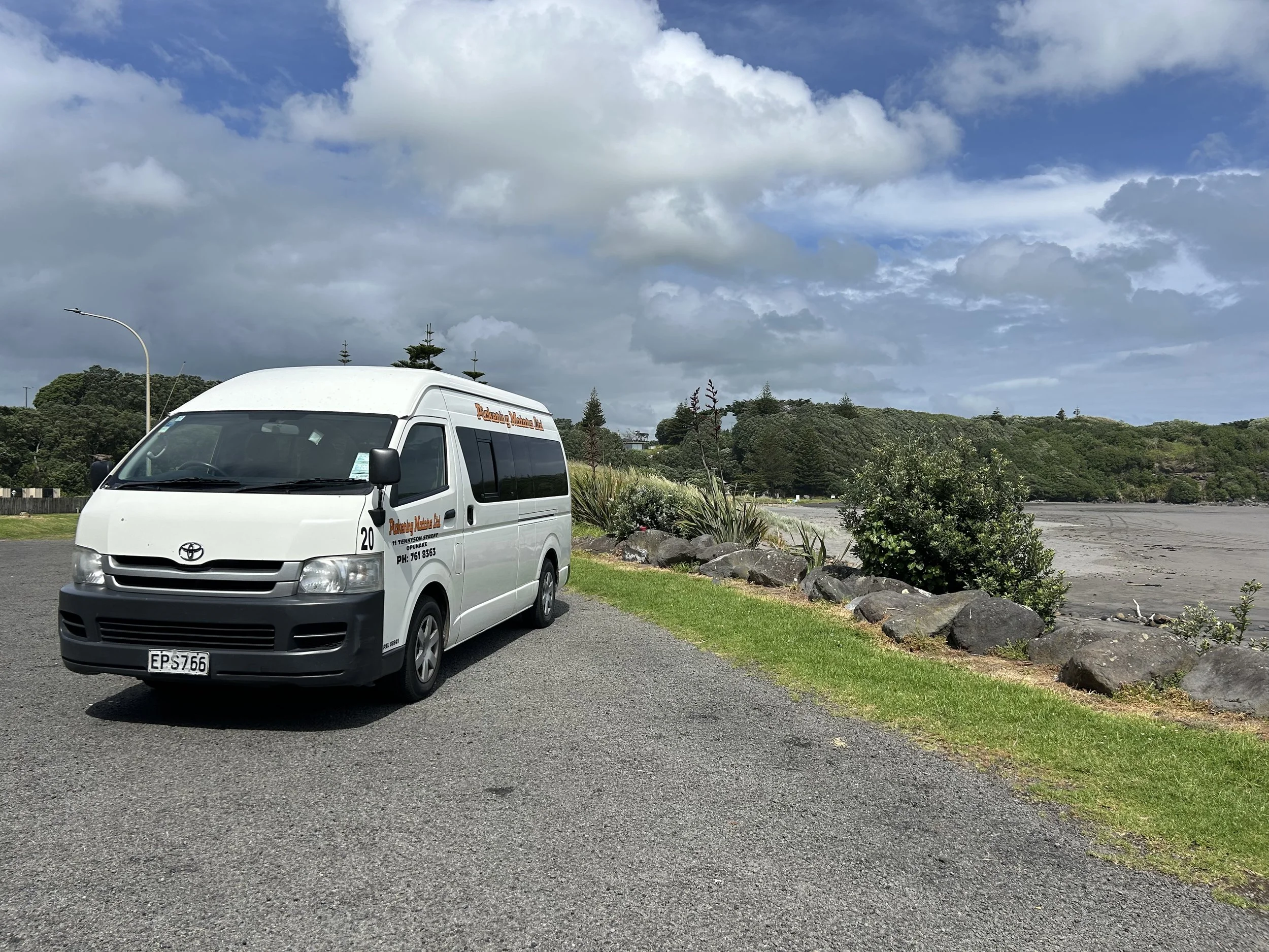 A white Toyota van parked on a gravel surface beside a coastal area with rocks, bushes, and green trees under a partly cloudy sky.