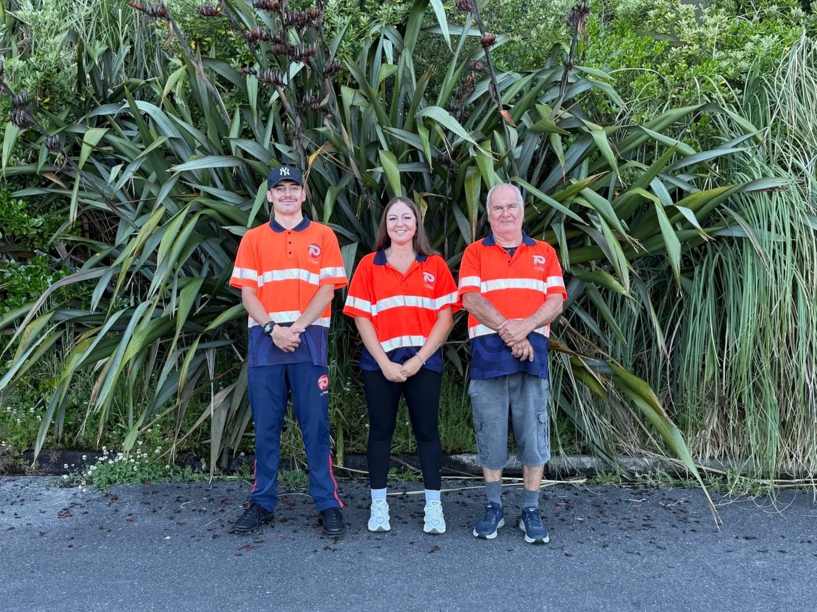 Three people standing in front of large leafy plants, wearing matching orange and navy uniforms.