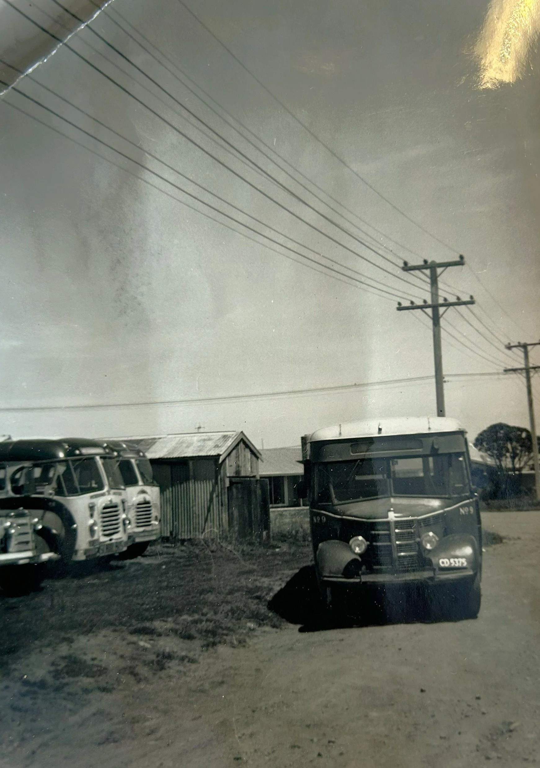 A black and white vintage photograph showing an old bus parked on a dirt street, with wooden houses and utility poles with power lines in the background.