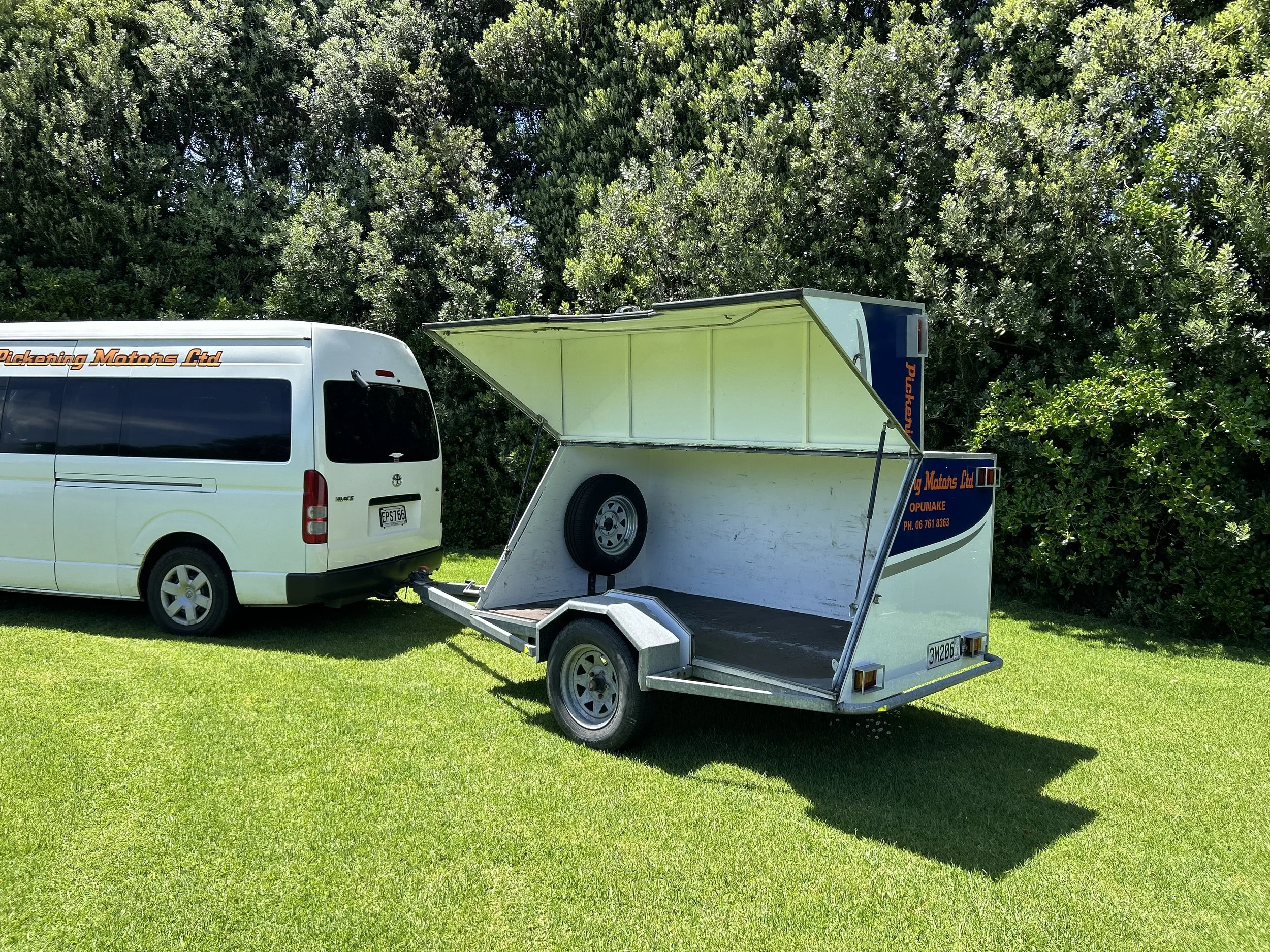 A white van with tinted windows and black trim attached to a trailer with an open side, showing a spare tire inside, parked on green grass in front of bushes.