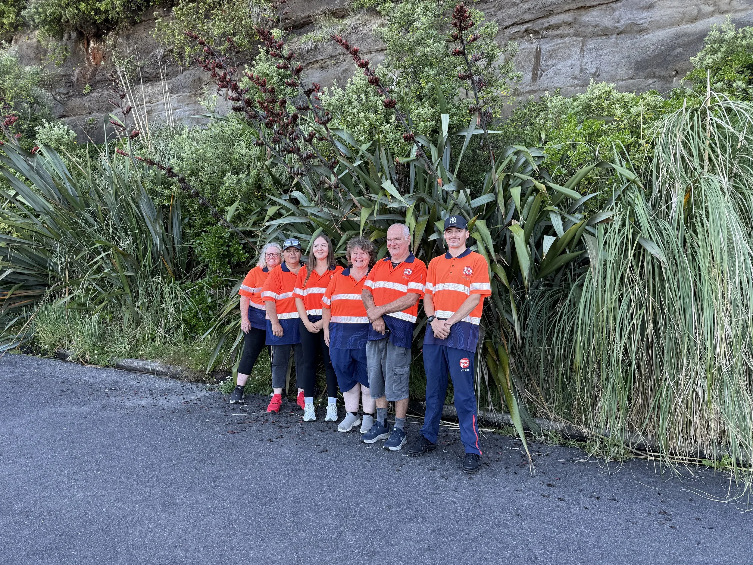 Group of six people standing together in front of large plants and a rock wall, all wearing matching orange and navy-blue uniforms.