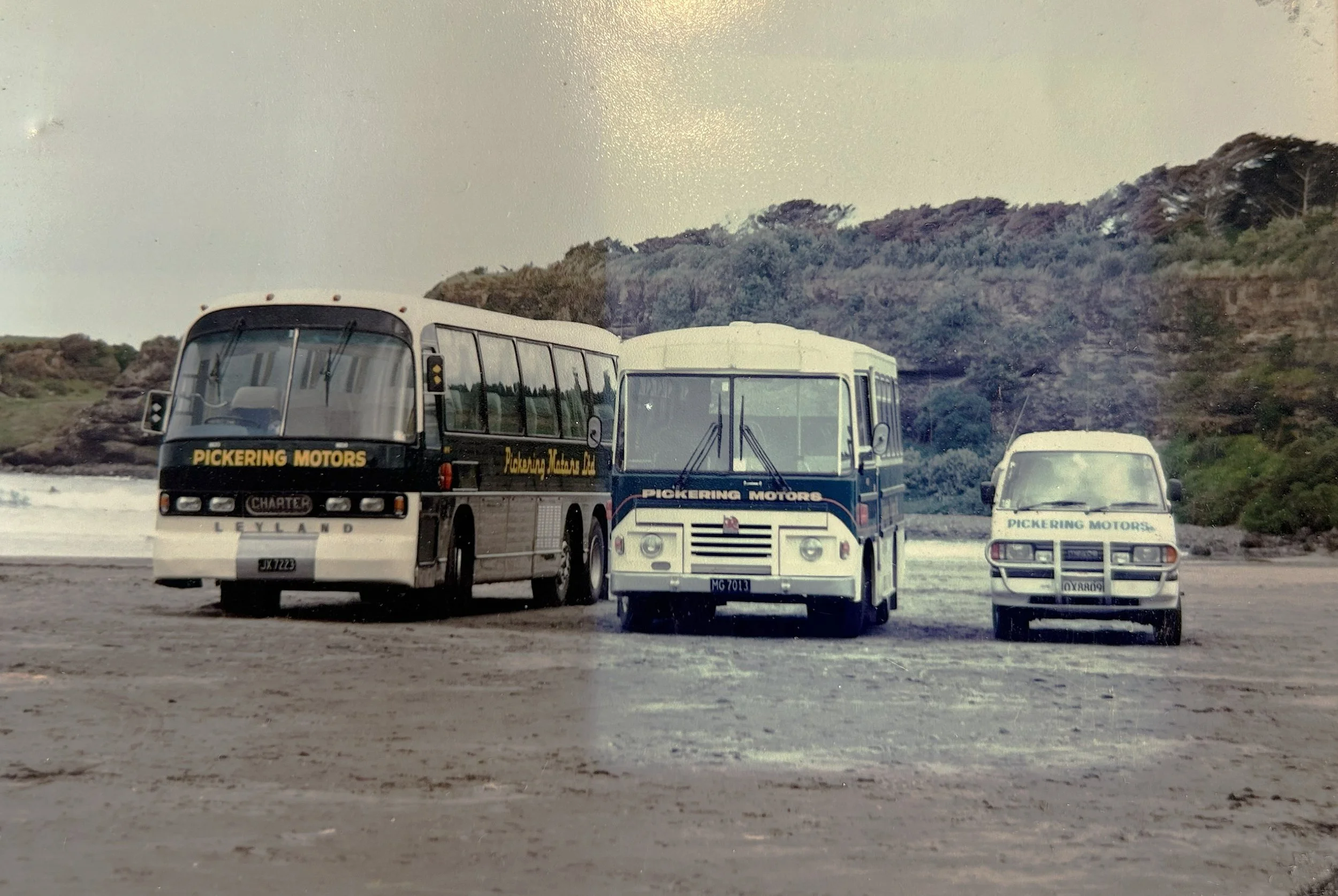 Three vintage buses with 'Pickering Motors' signage parked on a beach with rocky hills and water in the background.