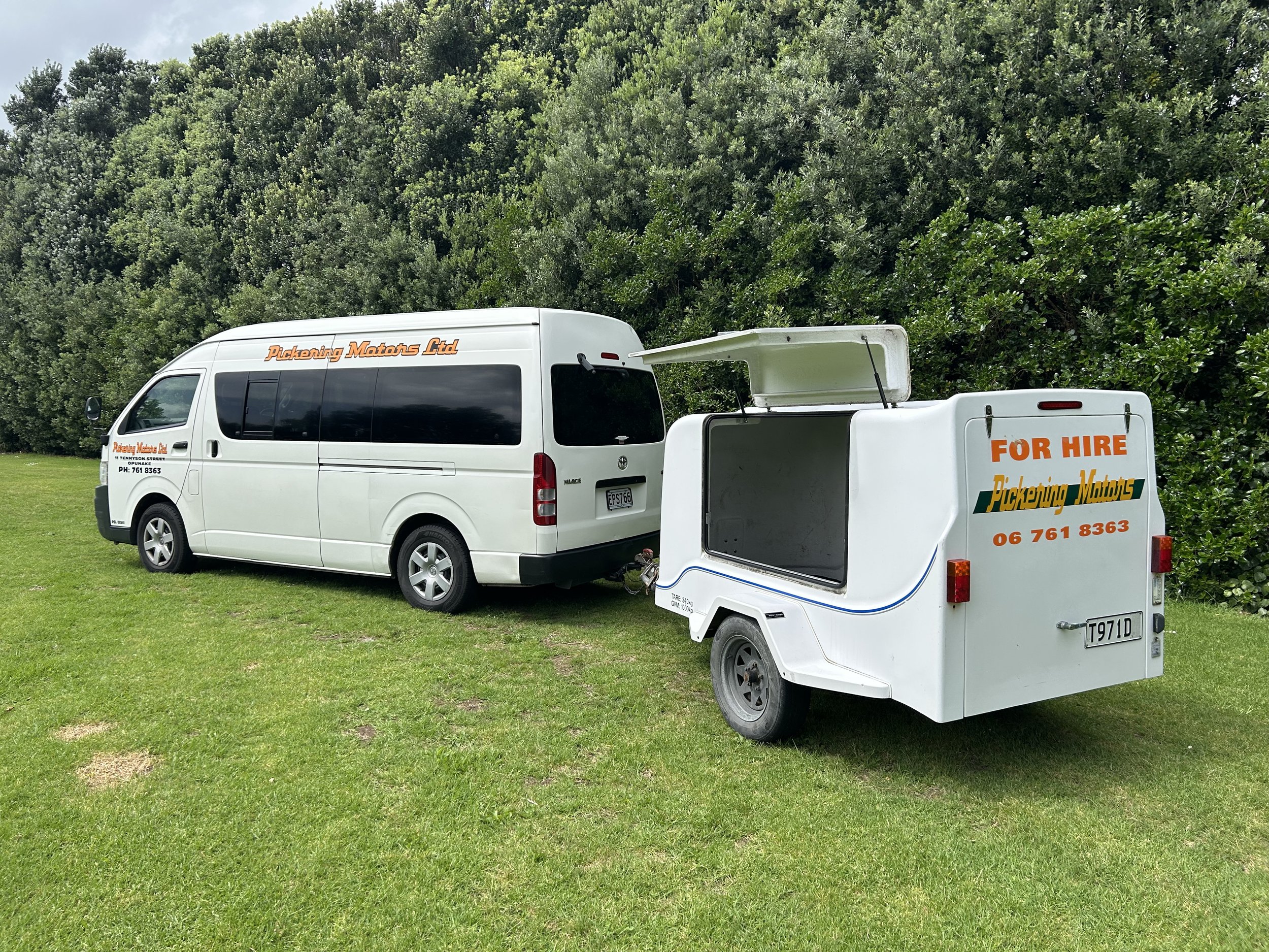 White van with black tinted windows and orange lettering parked on grass, attached to a small white food trailer with orange and green lettering that says 'For Hire' and 'Pickering Mowers,' with open storage at the top, set against green trees and bu