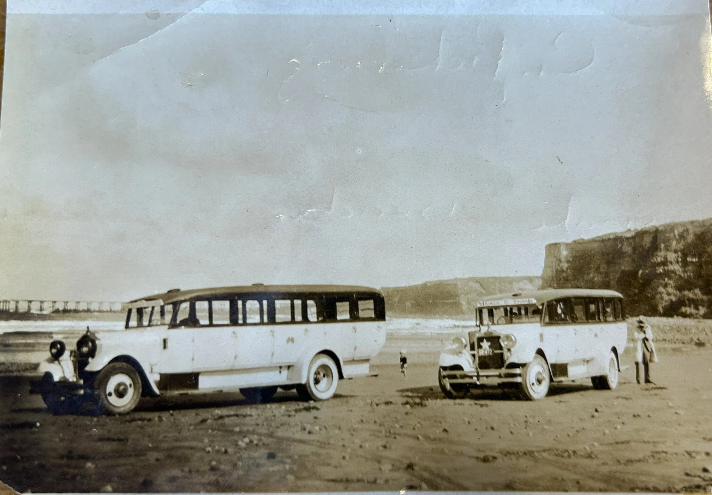 Two vintage buses parked on a beach with a cliff and ocean in the background. There are three people near the buses, including a woman with a hat.