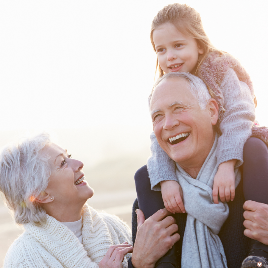 Grandfather with granddaughter on shoulders, smiling and looking at grandmother, outdoors, warm sunlight.