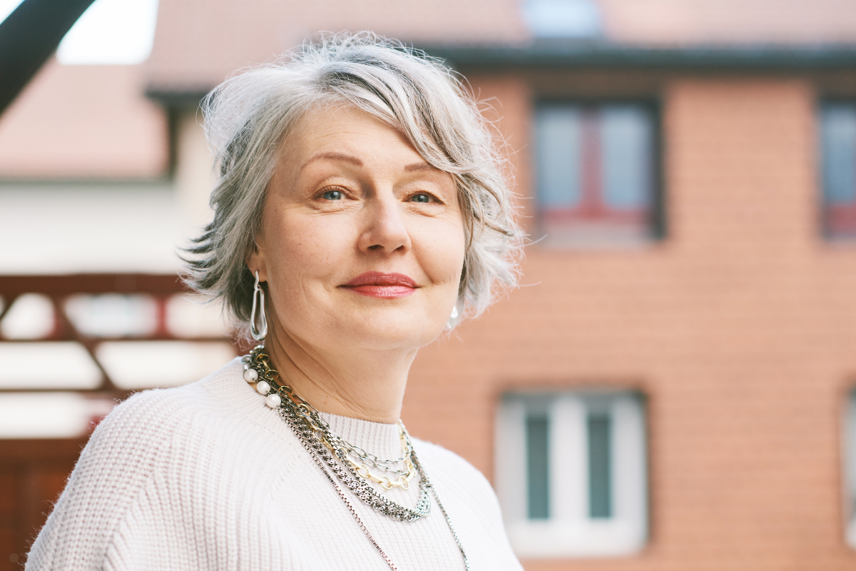 A portrait of an older woman with short gray hair, wearing a white knit sweater and layered necklaces, standing outdoors with a brick building and windows in the background.
