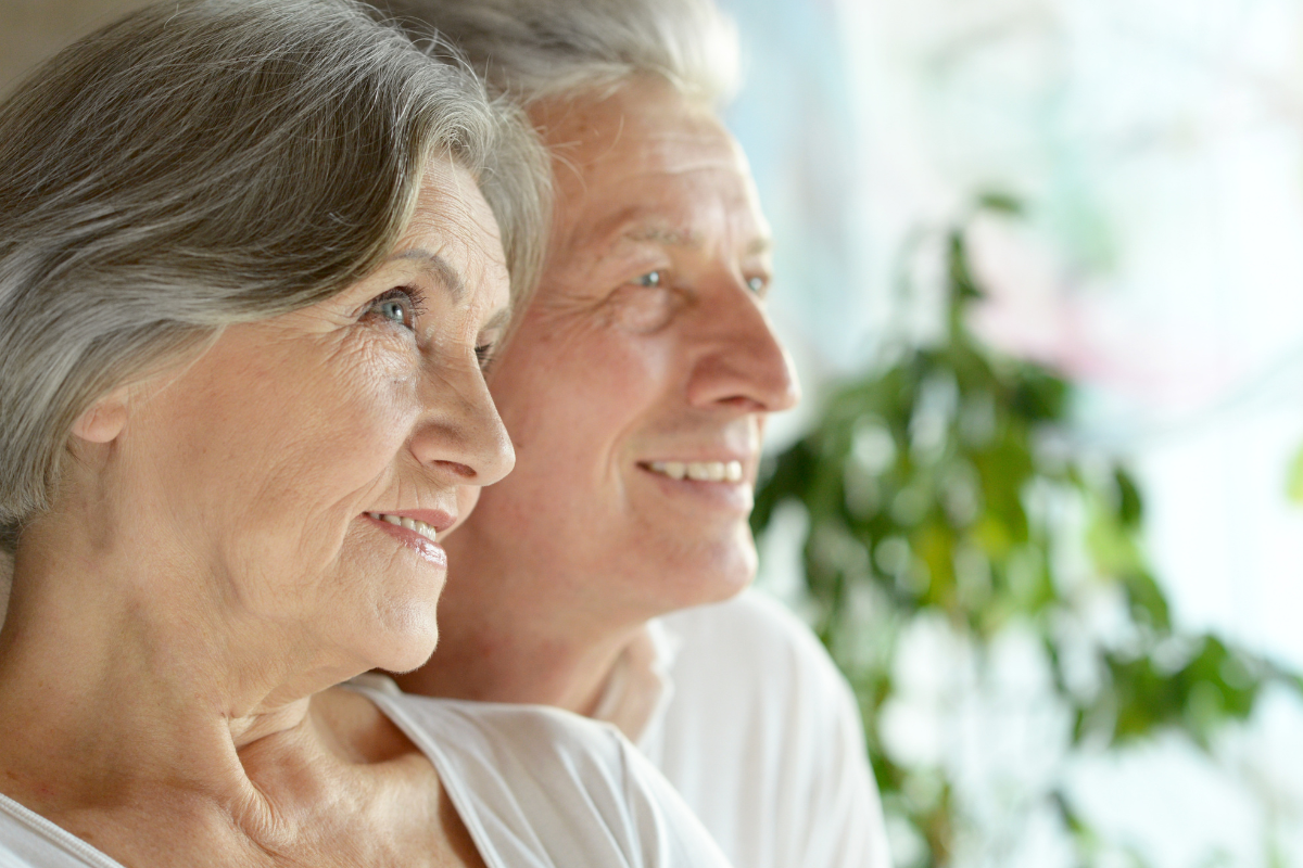 Close-up of an elderly couple smiling and looking to their right, with a blurred background of greenery and sunlight.