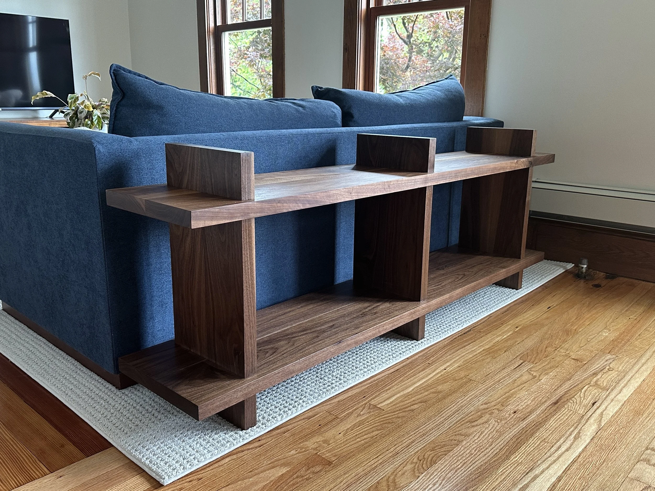 A living room with a dark blue sofa and a wooden shelving unit behind it, placed on a light-colored rug on wooden flooring, with windows showing trees outside.