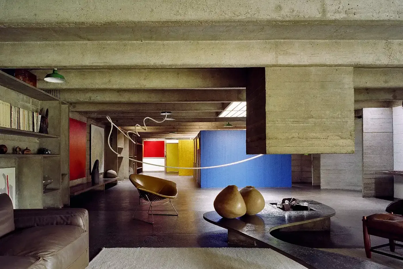 Sala modernista com paredes de cor cinza, em estilo brutalista brasileiro, móveis de época, cadeiras e objetos decorativos, teto de concreto exposto, iluminação natural e iluminação artificial