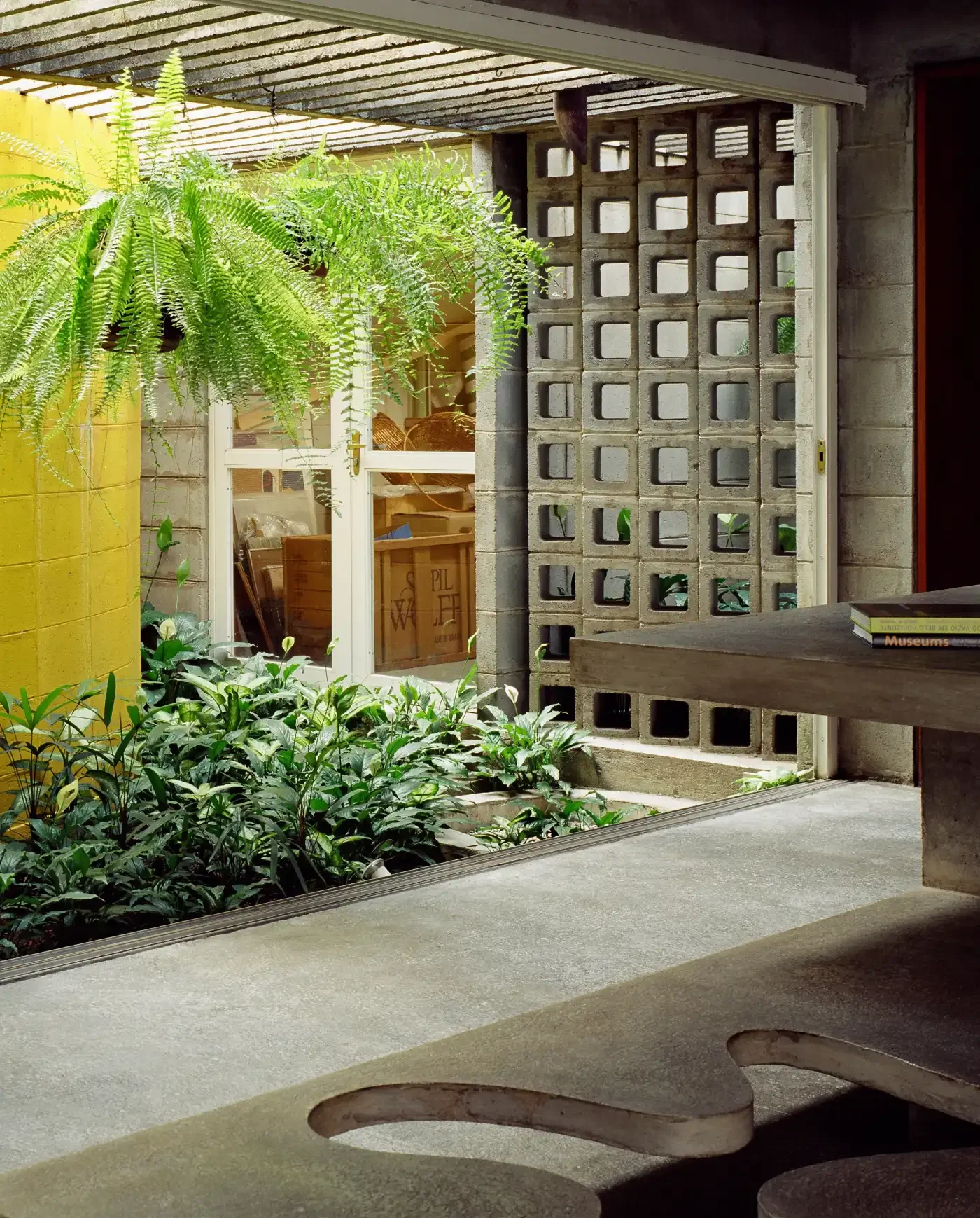 área de varanda com plantas verdes e uma parede de blocos de cimento, uma janela com portas de vidro e uma mesa de madeira com alguns livros