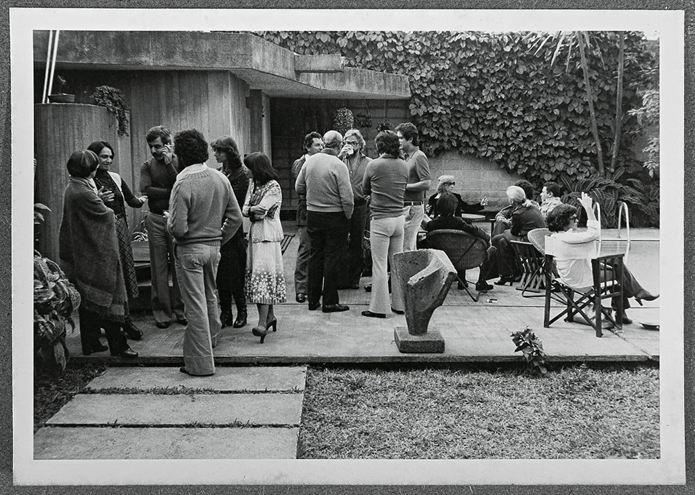 Grupo de pessoas conversando e socializando em um ambiente ao ar livre, com algumas sentadas e outras em pé, em um espaço de jardim ou terraço com plantas e decoração.